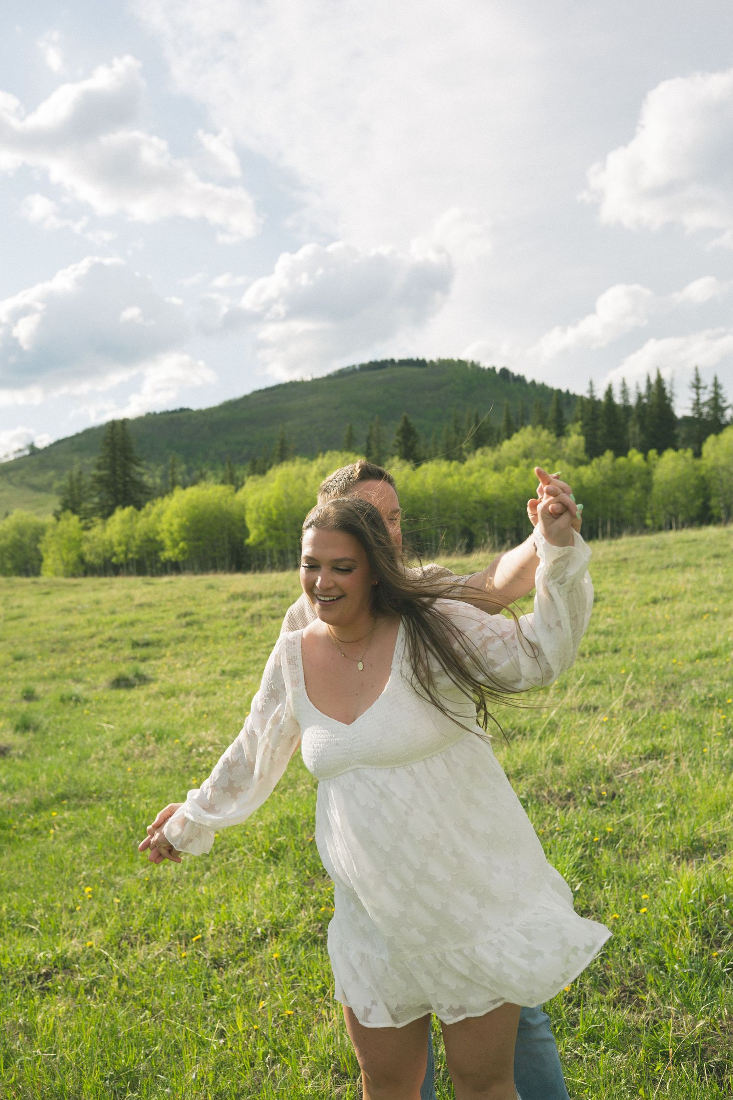 A person in a flowing white dress twirls carefree in a sunny mountain meadow surrounded by green hills.