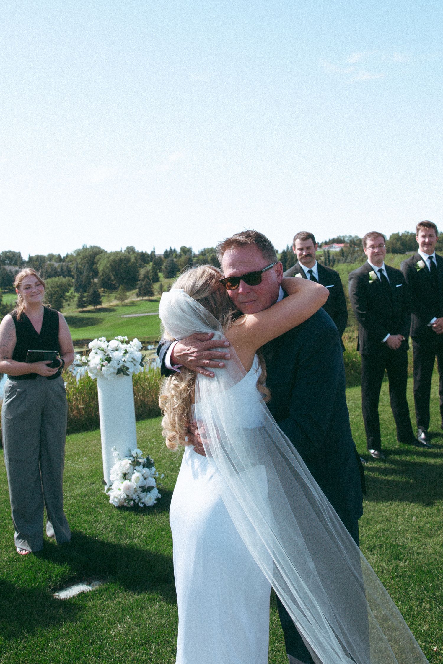 Emotional embrace during outdoor wedding ceremony with bridal party looking on.