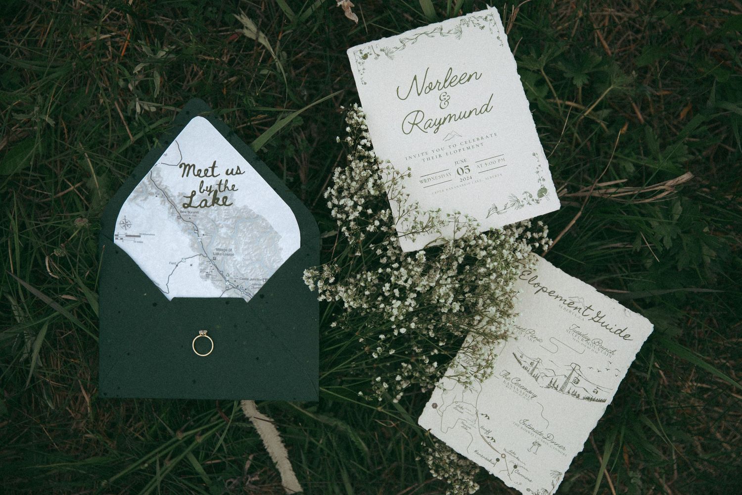 Elegant wedding invitations and green envelope with baby's breath flowers arranged on dark grass background.