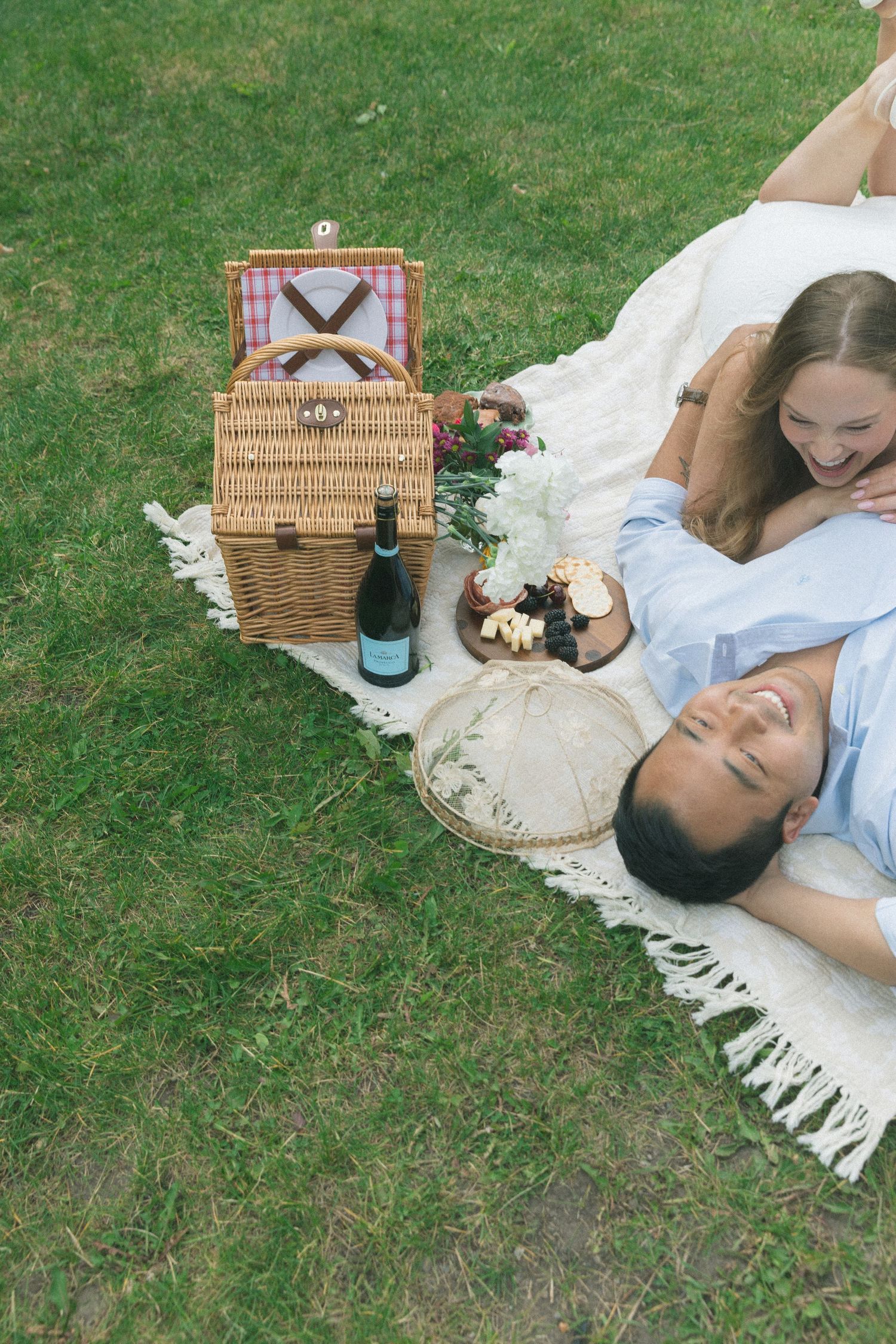 Couple enjoying a relaxing picnic on a grassy lawn with a wicker basket and white blanket spread out.