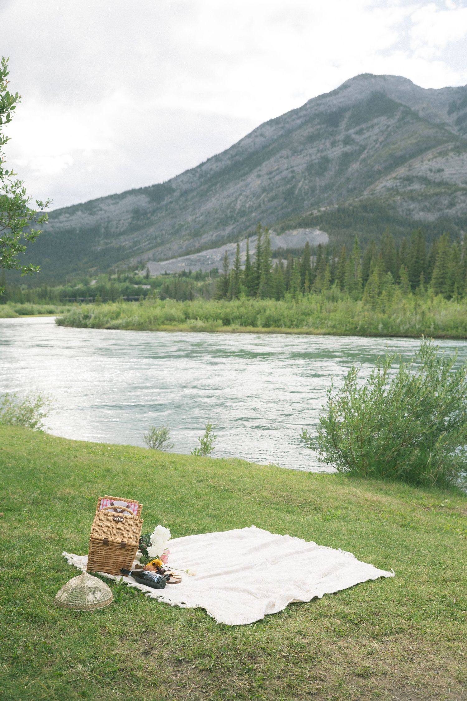 Picnic setup with white blanket and basket beside turquoise river with mountain backdrop.