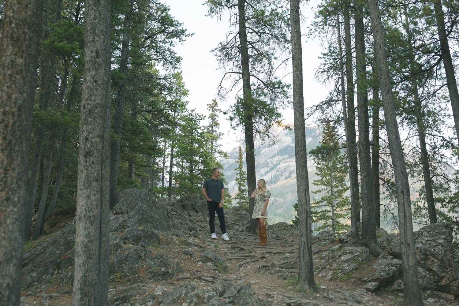 Two hikers explore a rocky forest trail surrounded by tall evergreen trees in misty mountain terrain.