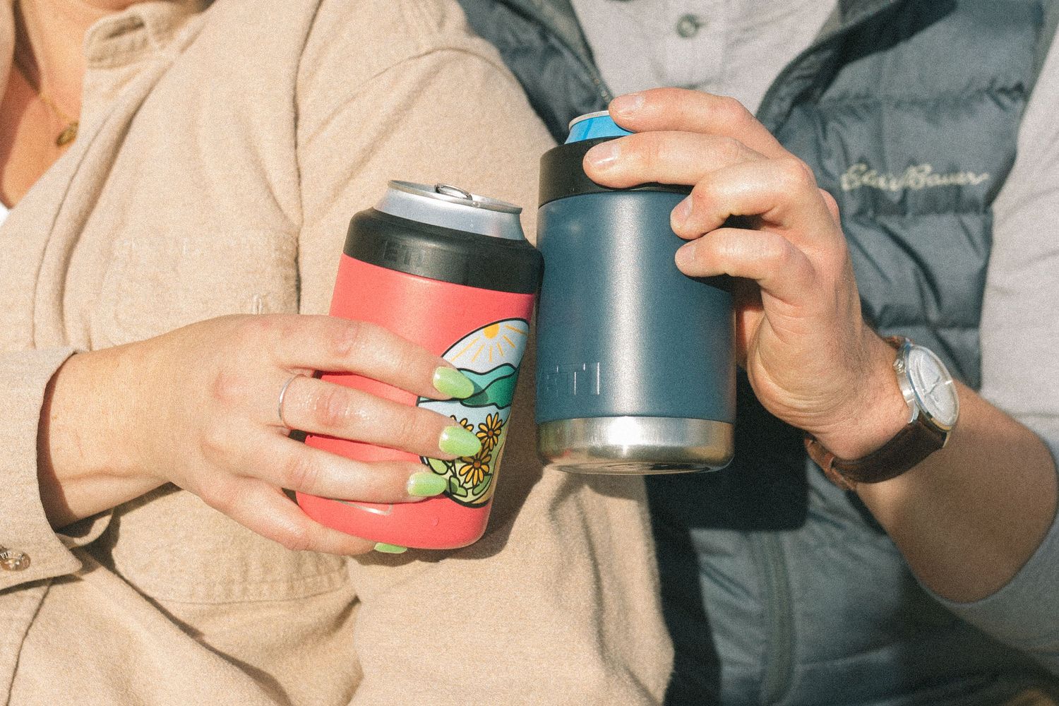 Two travel mugs being held together against mountain landscape backdrop.