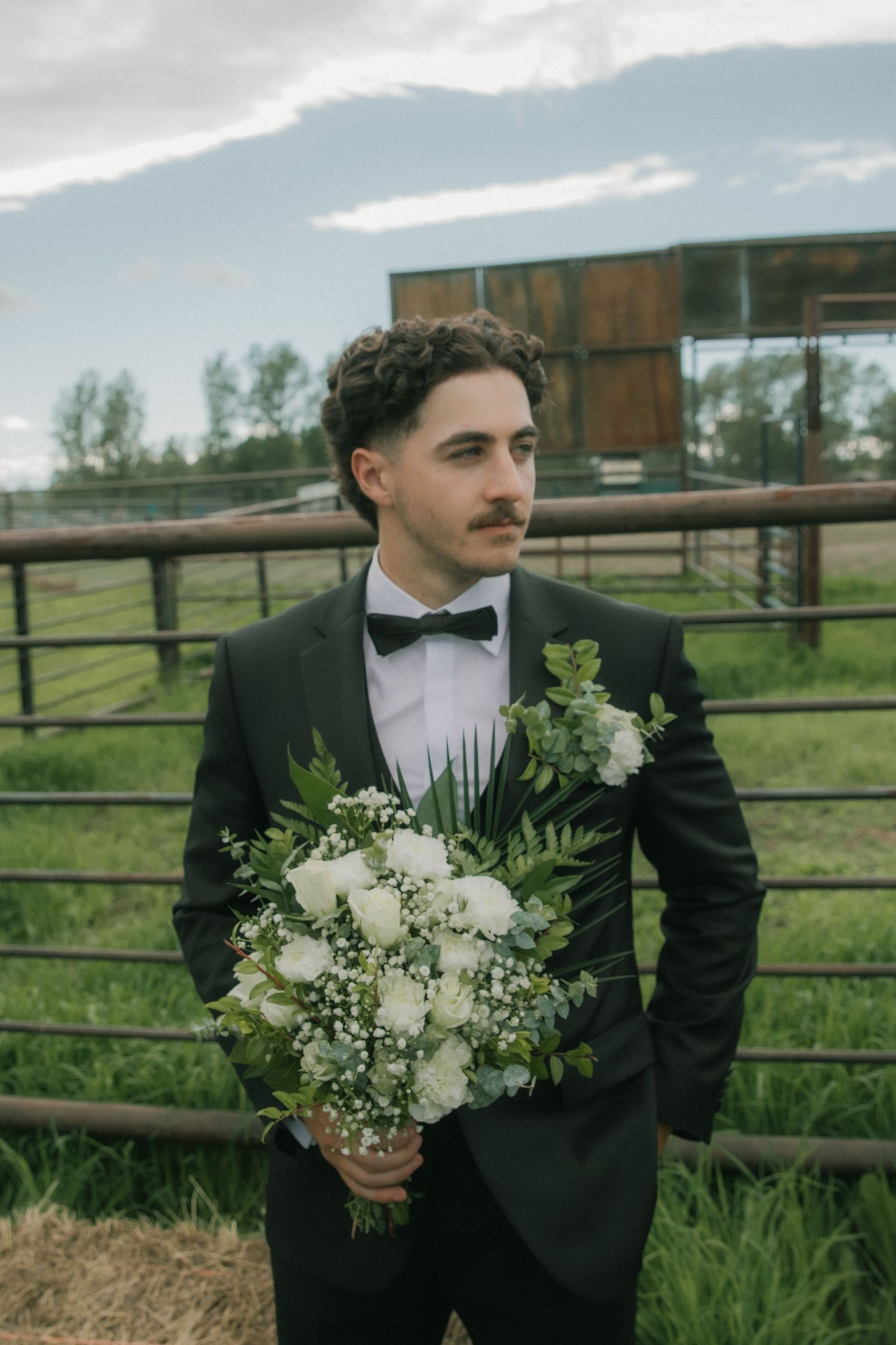 Dapper groom in black tuxedo holding white floral bouquet near farm fence outdoors.