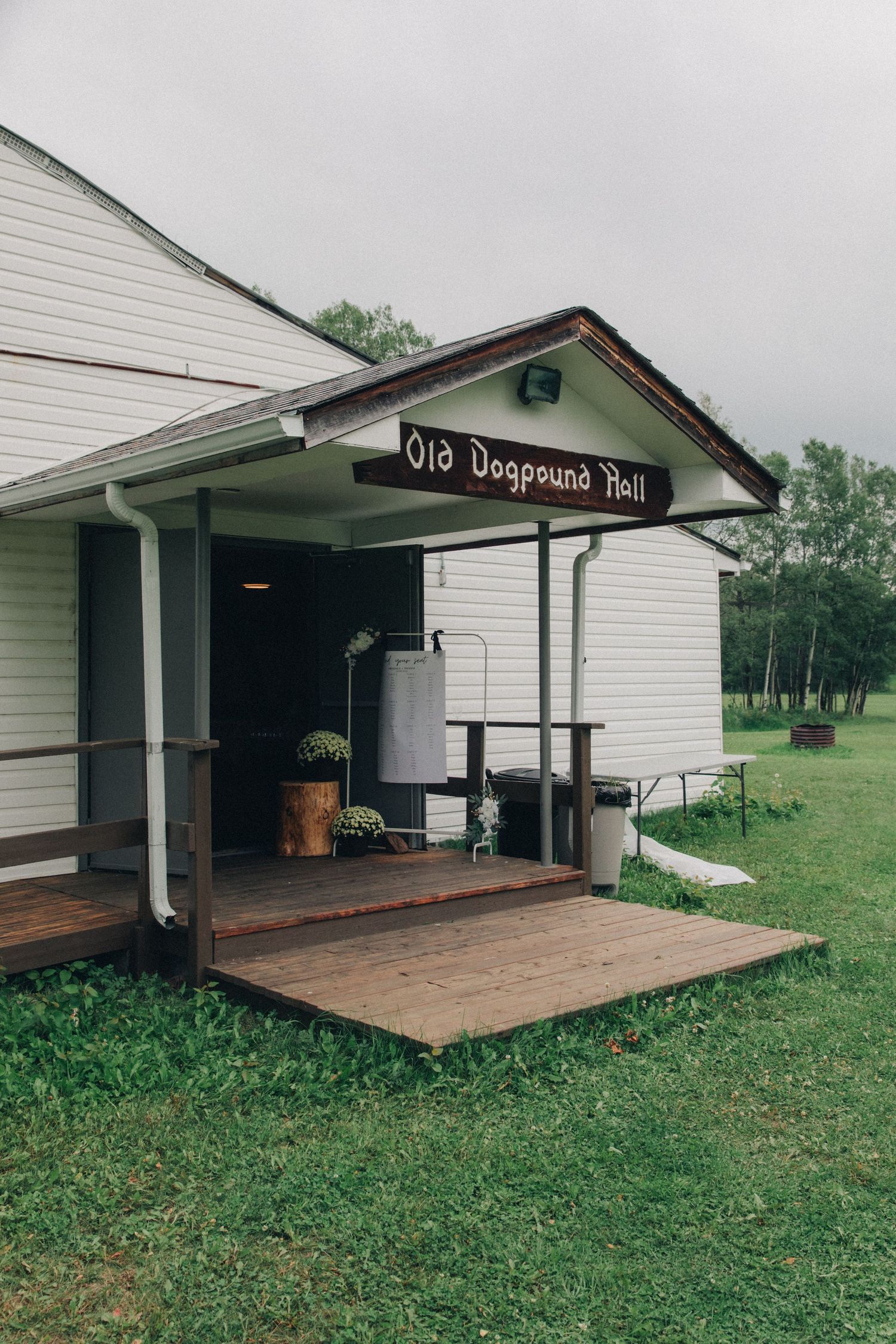 Rustic white wooden Old Dogpound Hall building with covered porch and wooden deck surrounded by green grass.