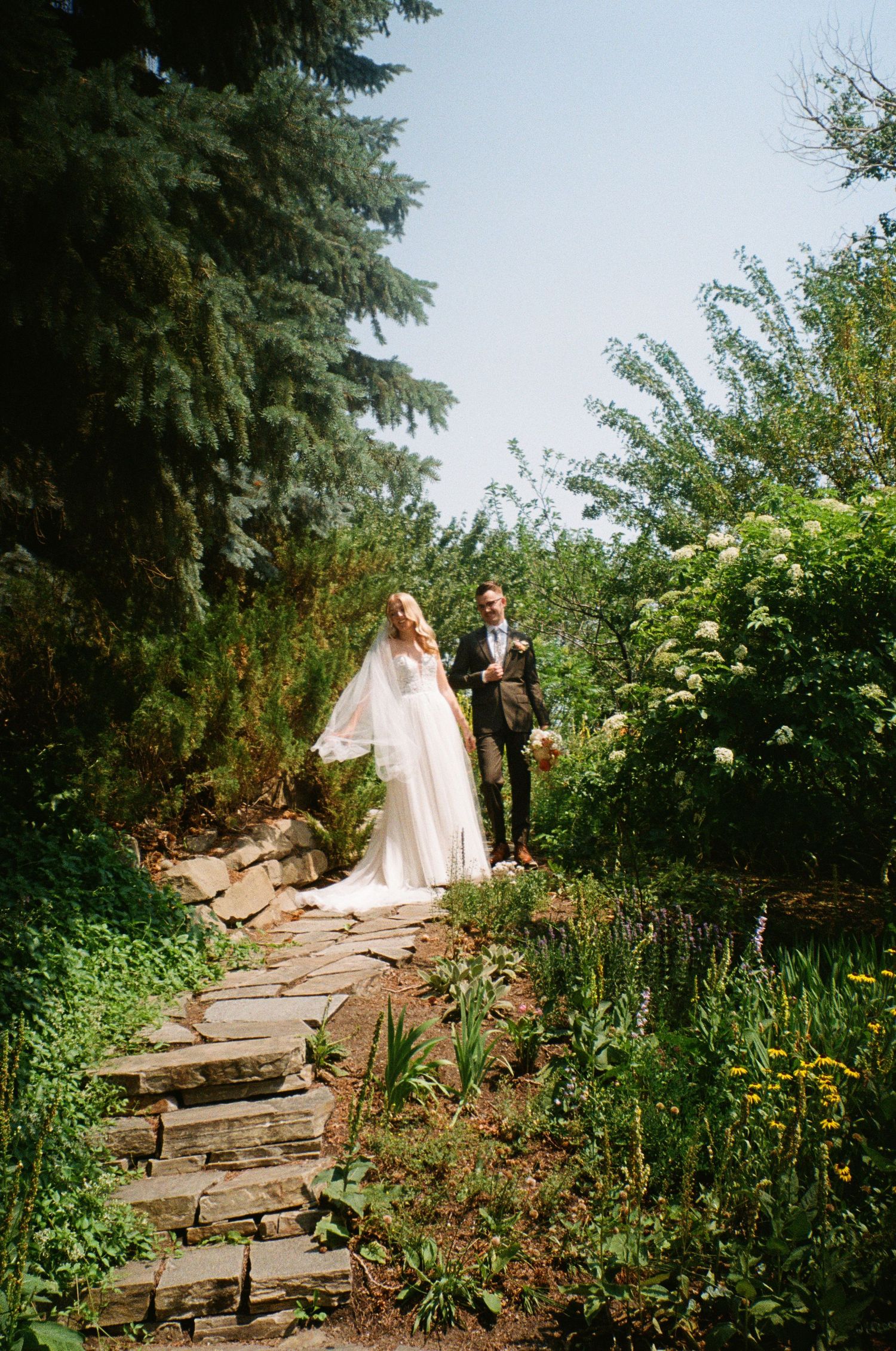 A couple on their wedding day poses on a rustic stone garden path surrounded by lush greenery and flowers.