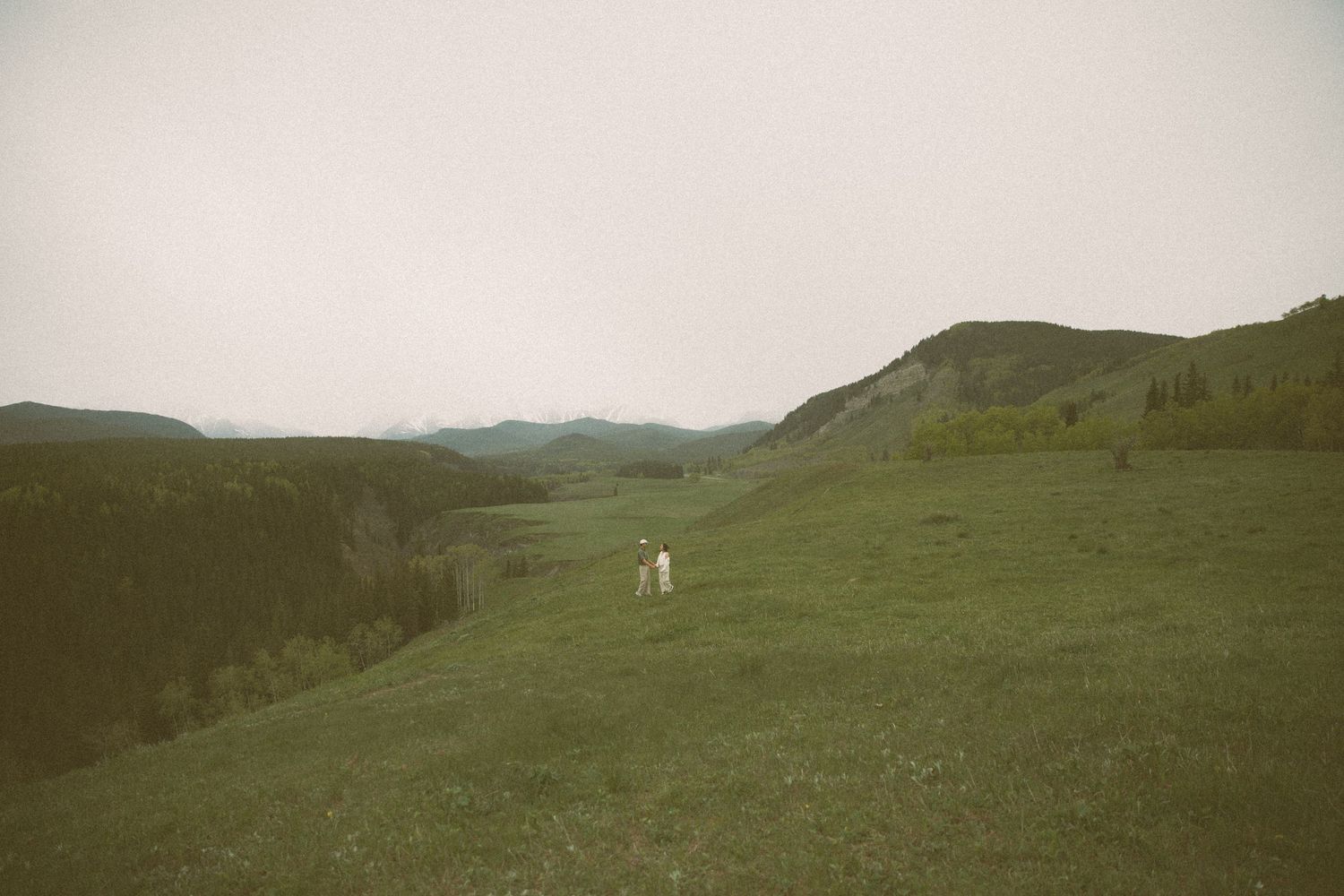 A distant figure in white walks across a remote green hillside landscape under an overcast sky.
