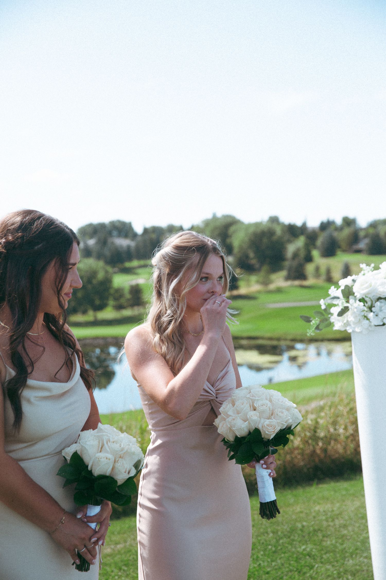 Bridesmaids in elegant dresses hold white bouquets at outdoor wedding ceremony.