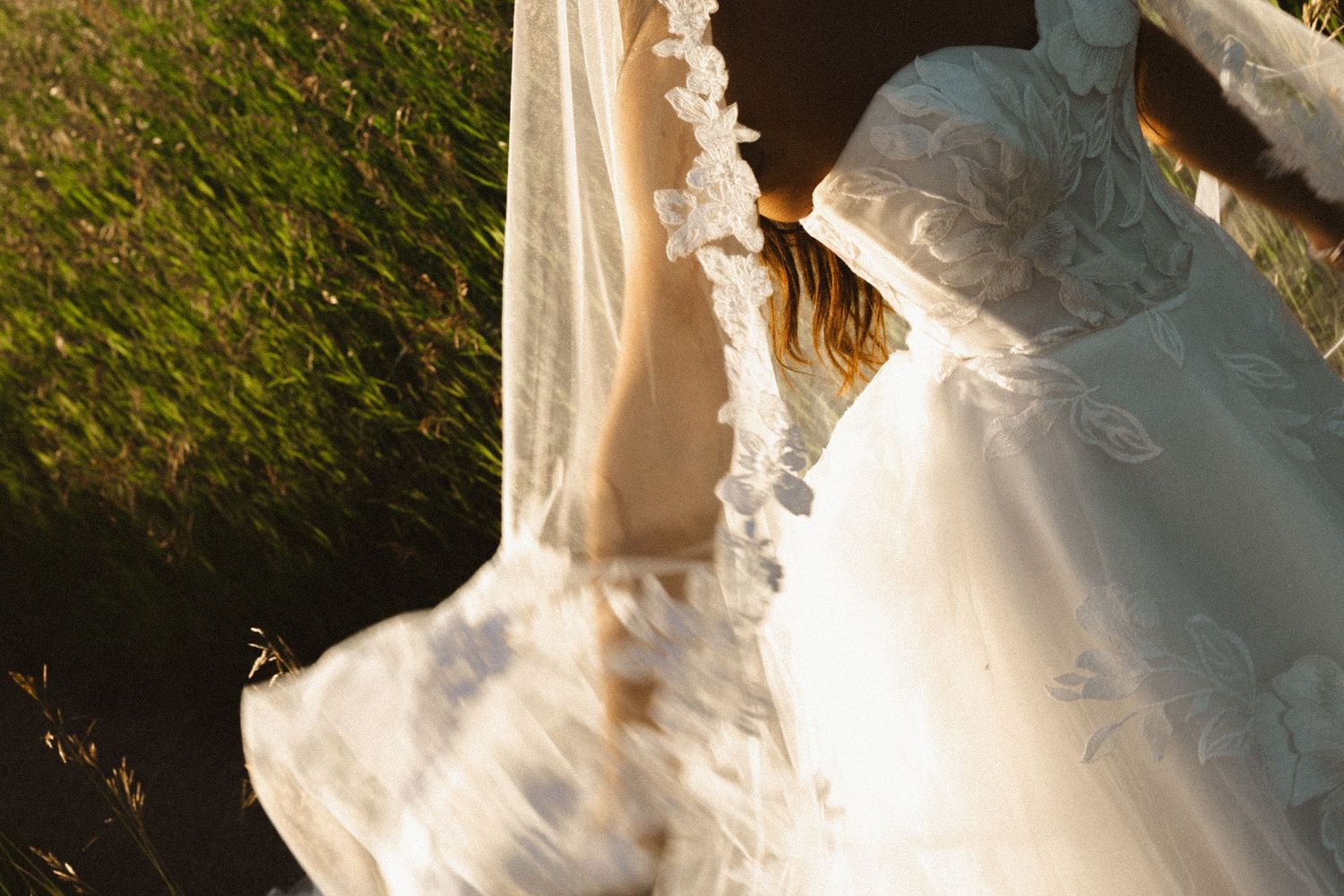 Bride in a flowing white wedding dress and floral veil captured in golden sunlight outdoors.