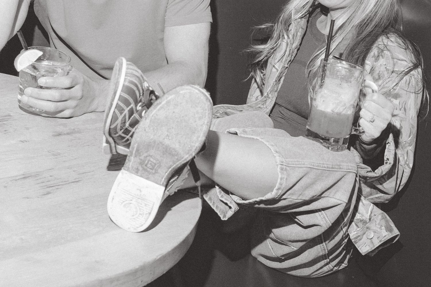 Black and white photo of two people sitting at a table with drinks, one person resting feet up showing shoe soles.