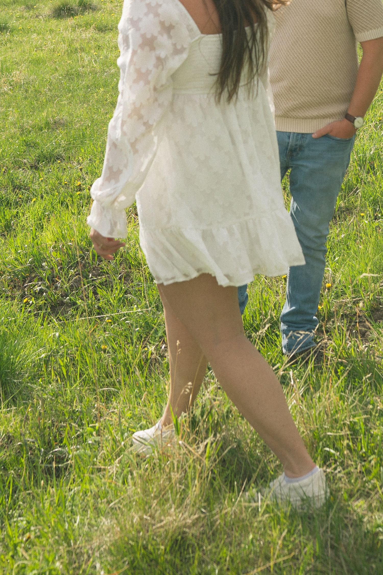A person wearing a white lace dress and sneakers stands in sunny green grass next to someone in jeans.