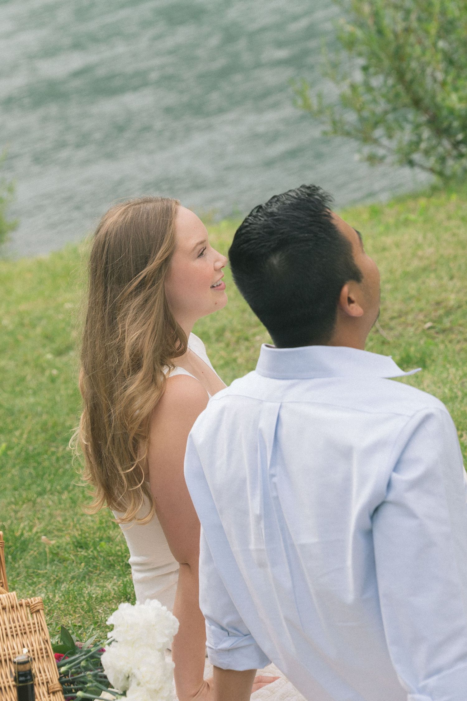 A couple shares a romantic moment on grass near water during a picnic with a wicker basket visible.