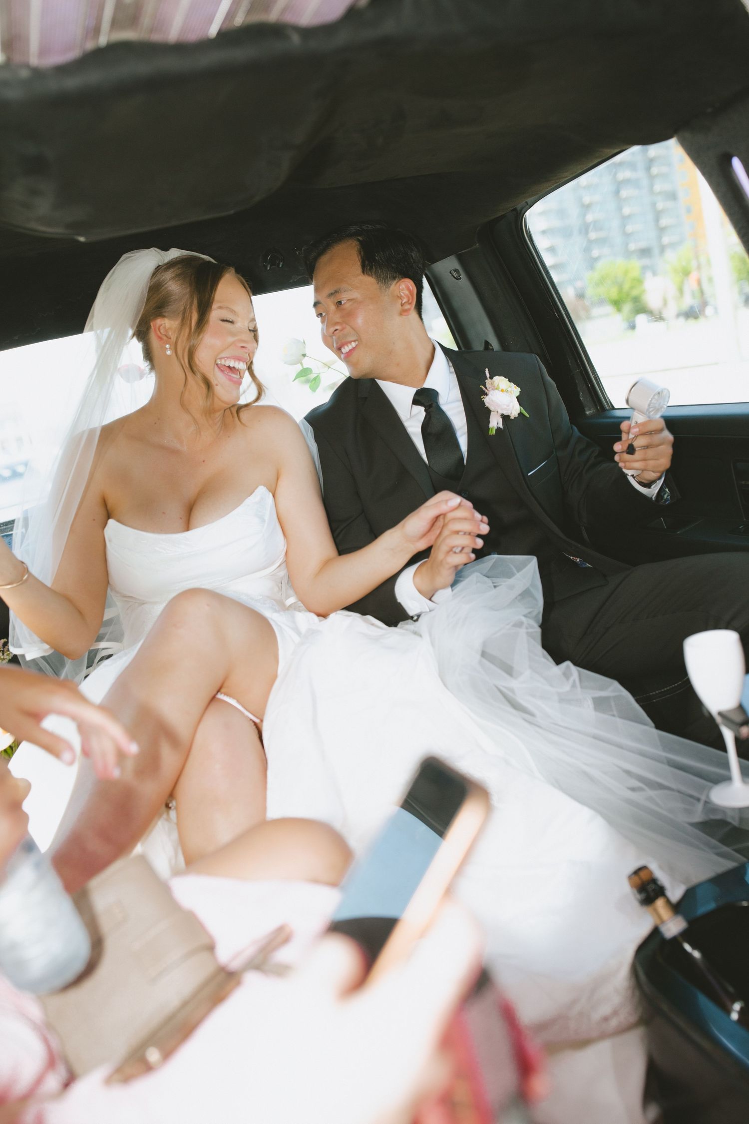 Happy newlyweds holding hands and champagne in a limousine, bride in white gown with veil, groom in black tuxedo.