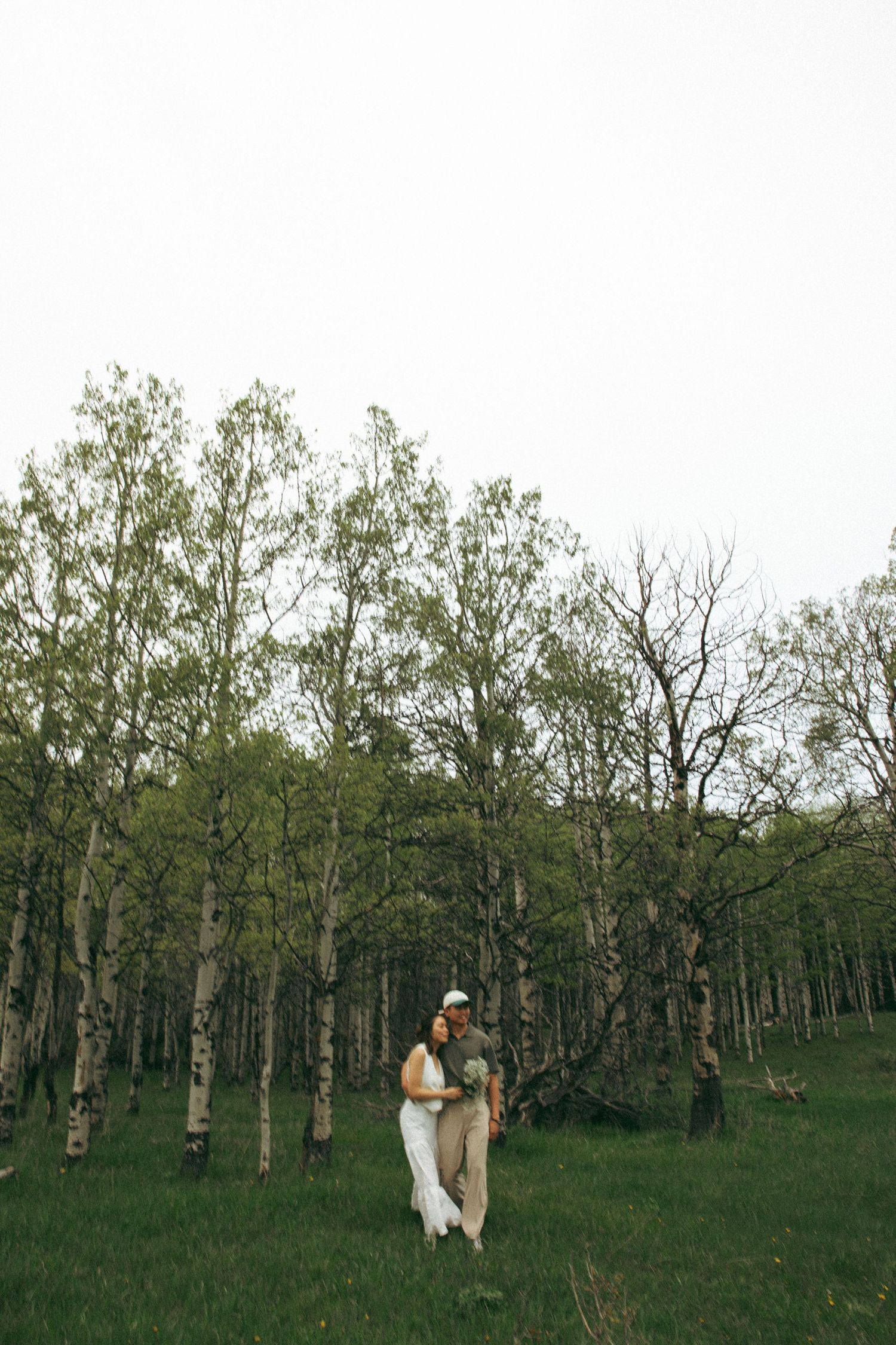 A couple in light-colored attire stands together among a grove of tall birch trees in a green field.