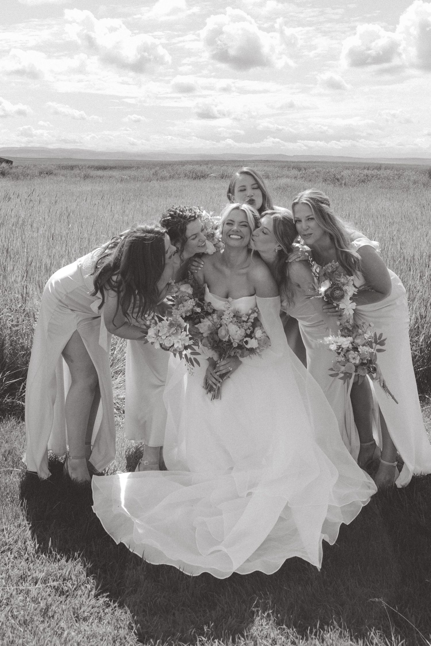 Bridesmaids in white dresses pose playfully in a field under cloudy skies.