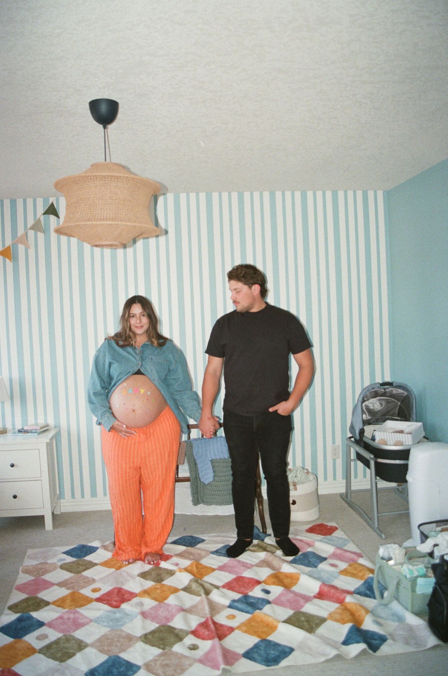 Two people stand in a room with blue striped wallpaper and a colorful patchwork quilt laid out on the floor.