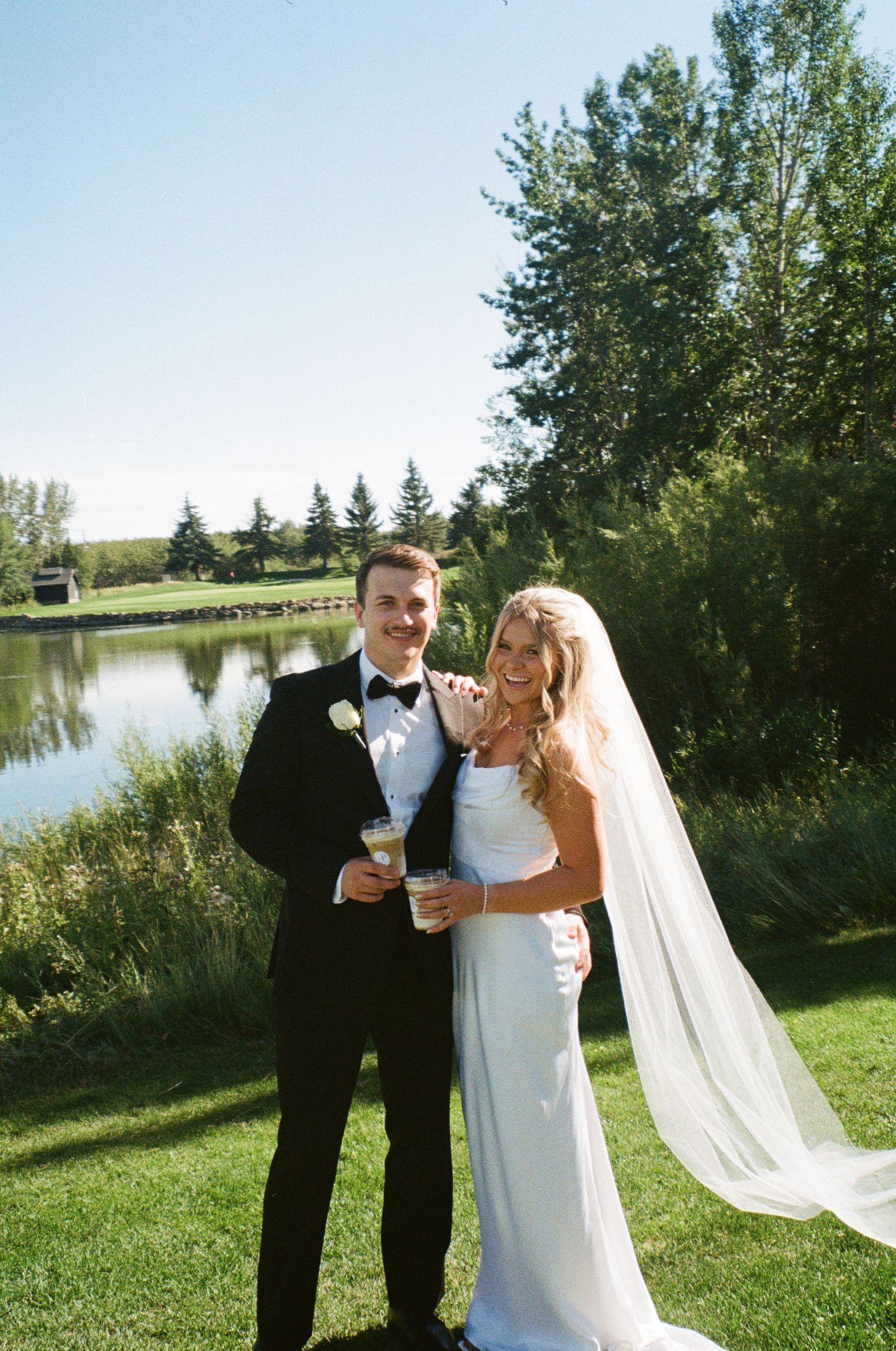 Newlyweds pose together on their wedding day by a serene lake with trees in the background.
