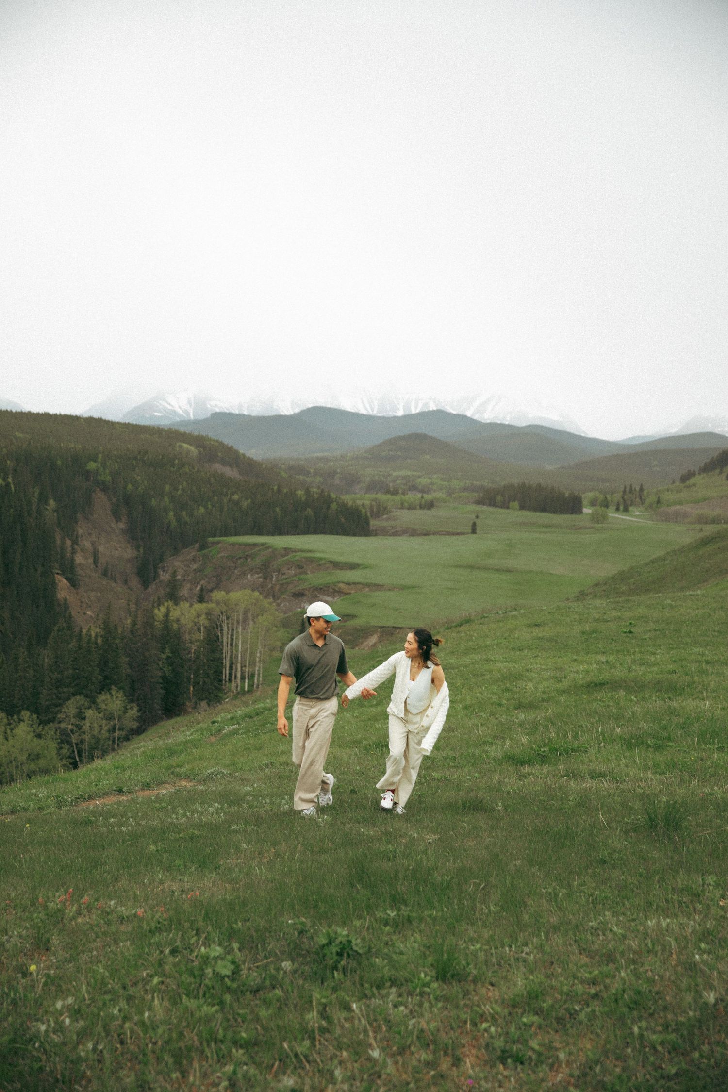 A couple walks hand in hand across a green mountain meadow with rolling hills in the background.