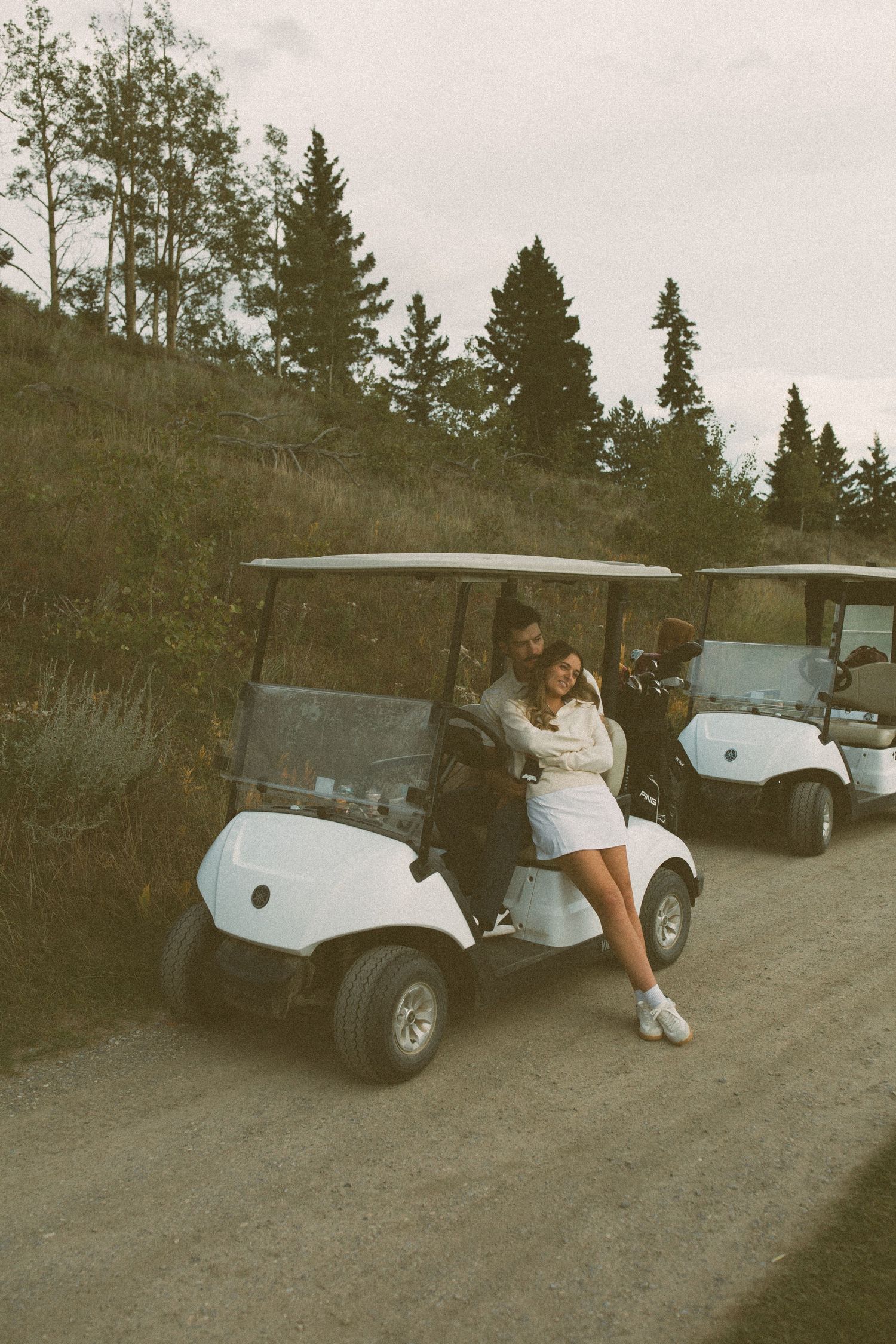 Couple posing with golf cart on hillside path with pine trees in background.