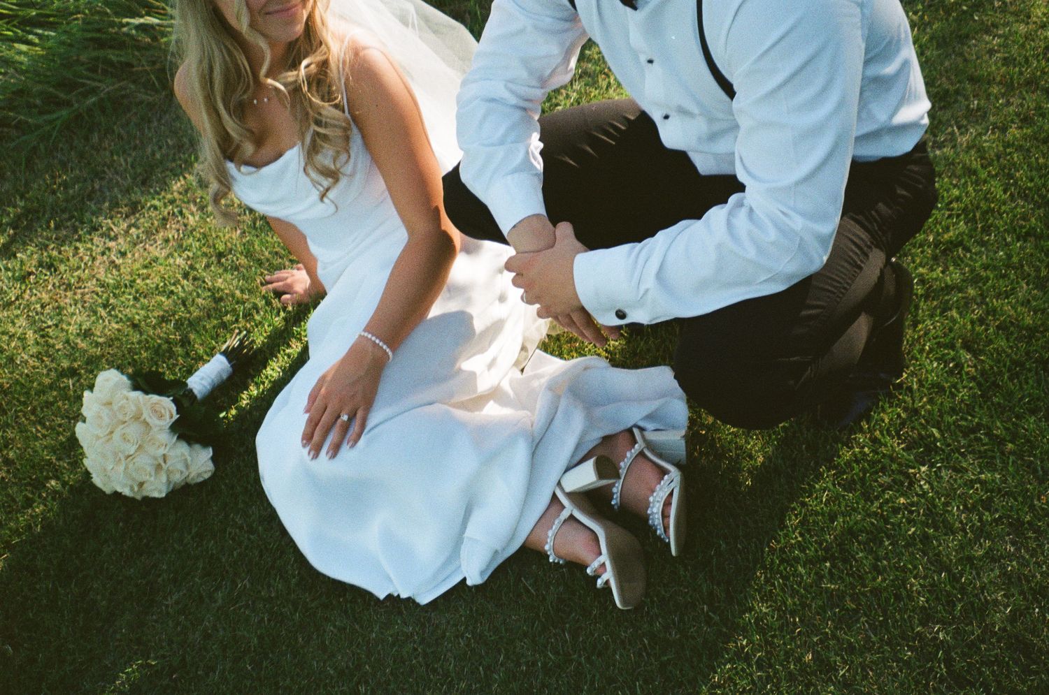 A couple sits on grass during their wedding, with a white bouquet and elegant shoes visible on the lawn.