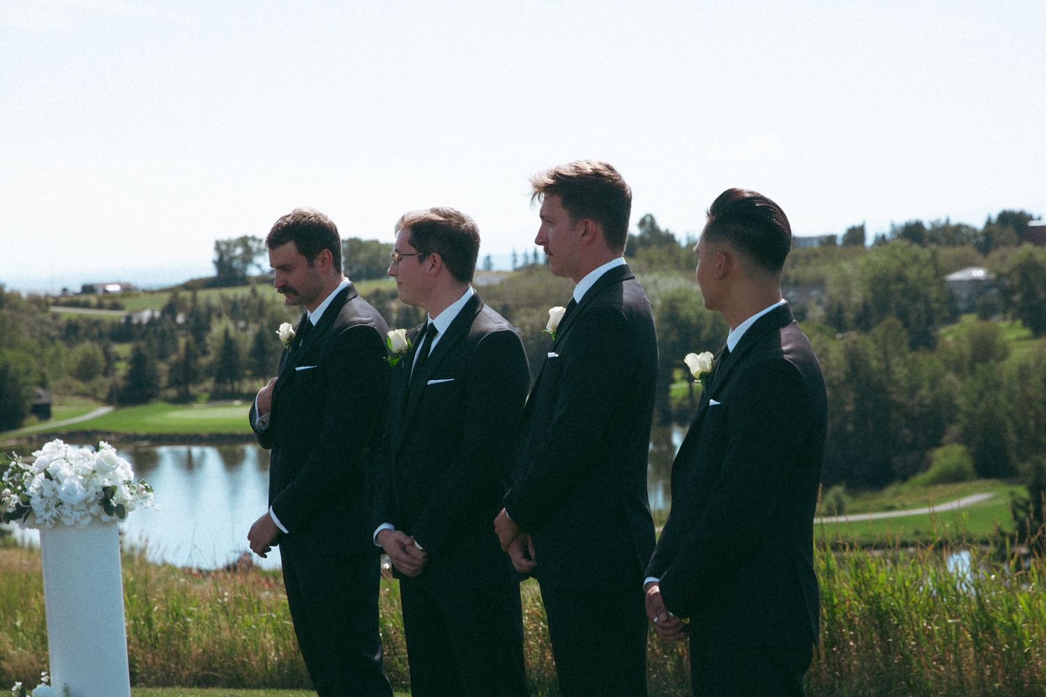 Wedding party in formal attire stands at scenic outdoor venue overlooking water.