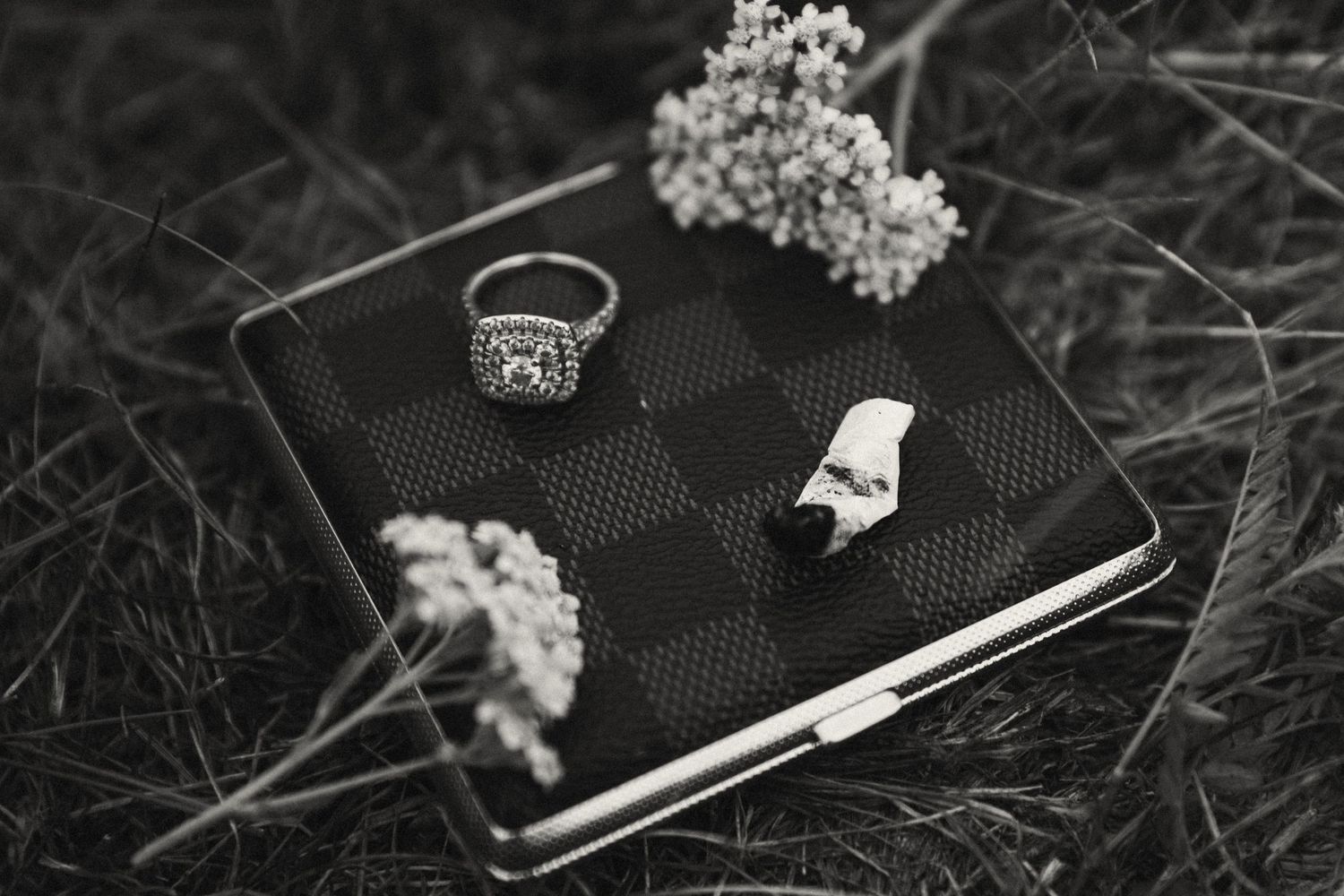 A black designer wallet with a ring and small white flowers arranged on grass in black and white photography.