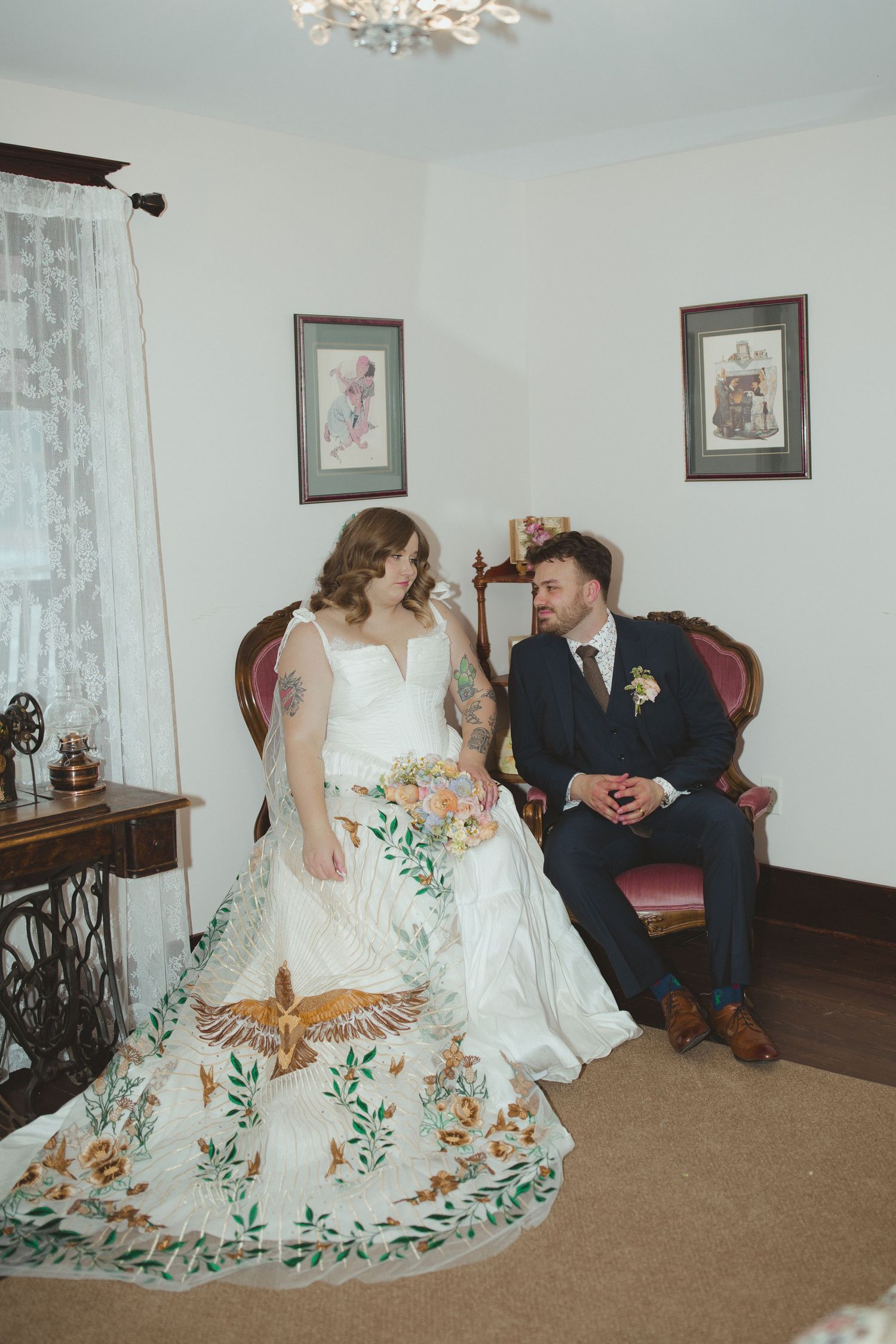 Bride in floral embroidered wedding gown holding bouquet stands beside seated groom in navy suit in vintage decorated room.