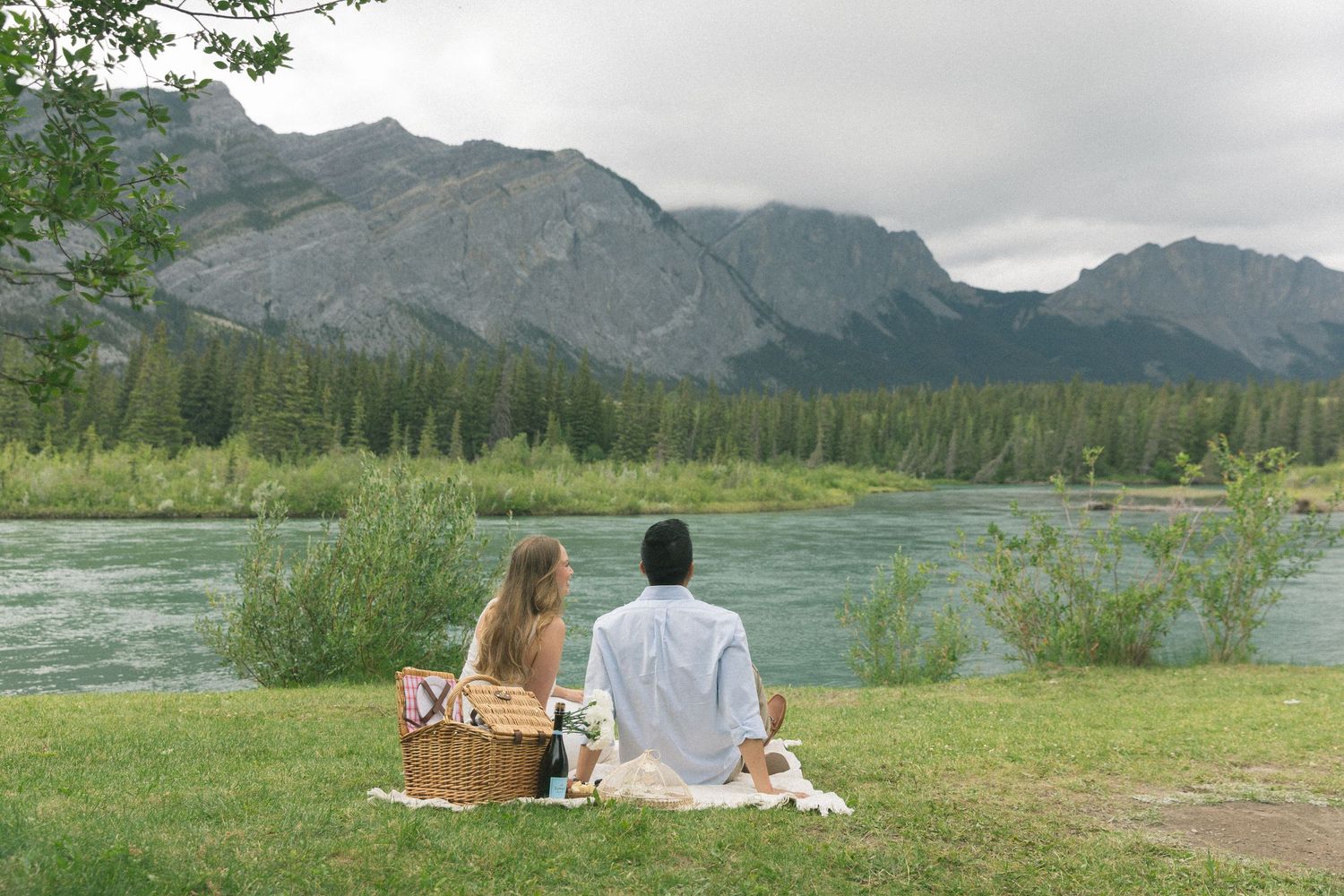 A riverside picnic scene in front of majestic mountains with evergreen forest.
