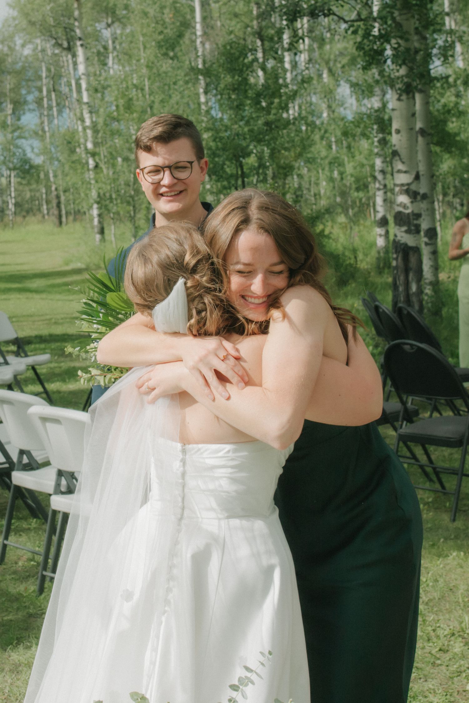 Bride in white gown hugging bridesmaids in green dresses outdoors among birch trees at wedding.
