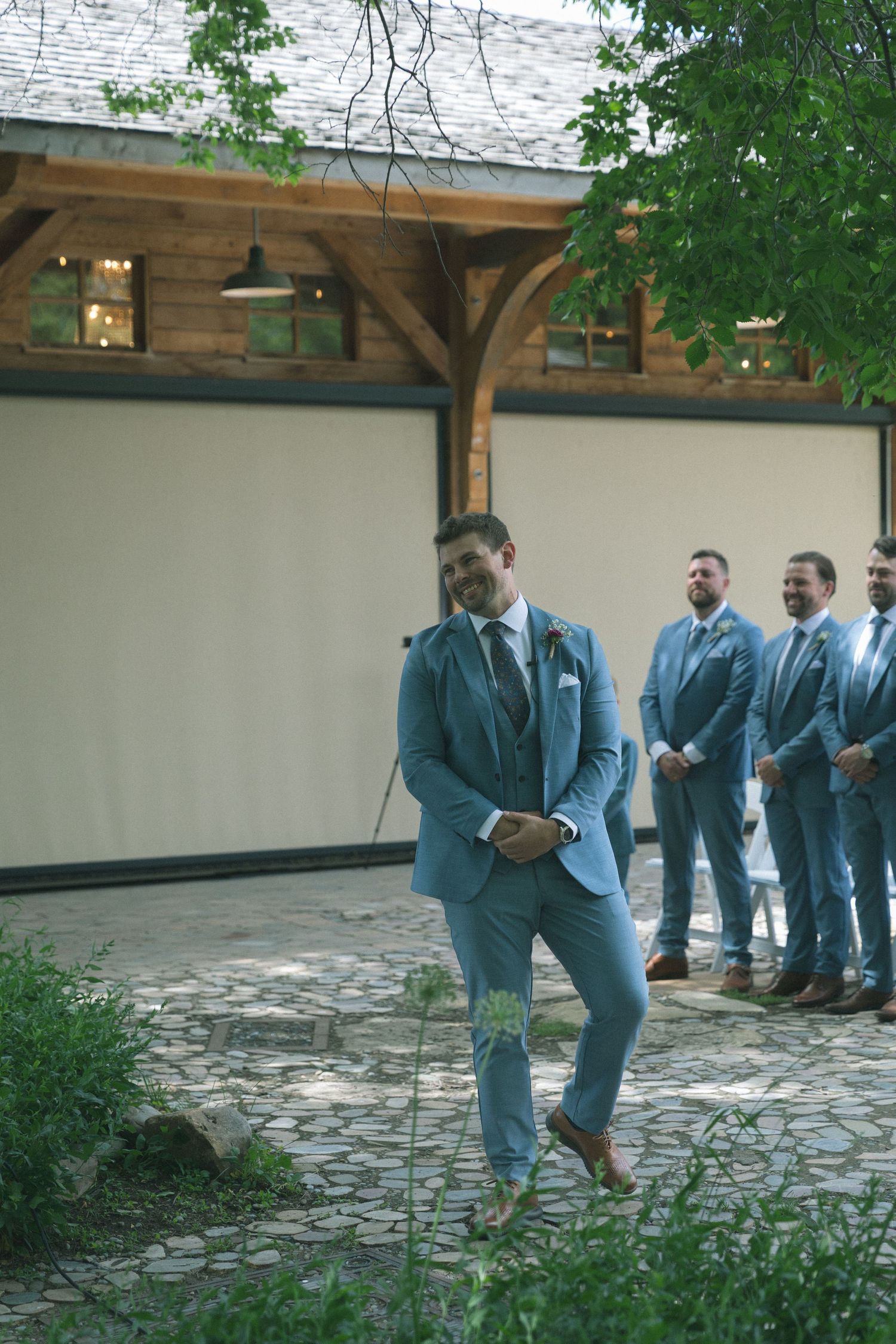 Groomsmen in light blue suits gather outside in a garden setting during a wedding celebration.
