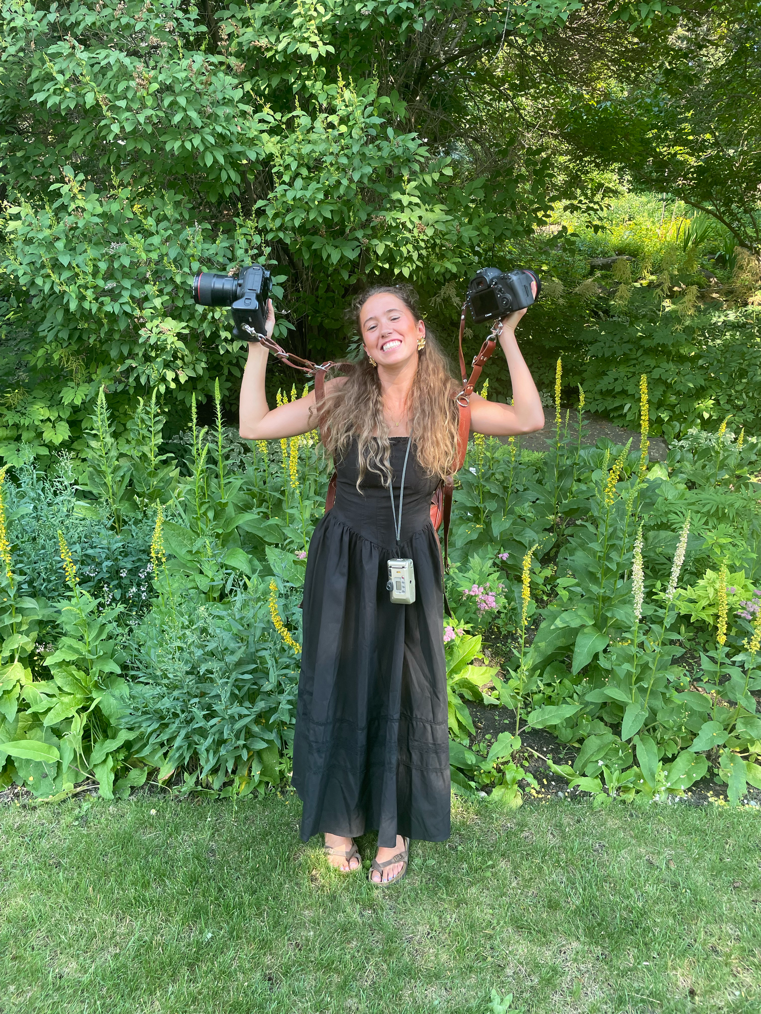 Wedding photographer in black dress holding two cameras triumphantly in garden setting.