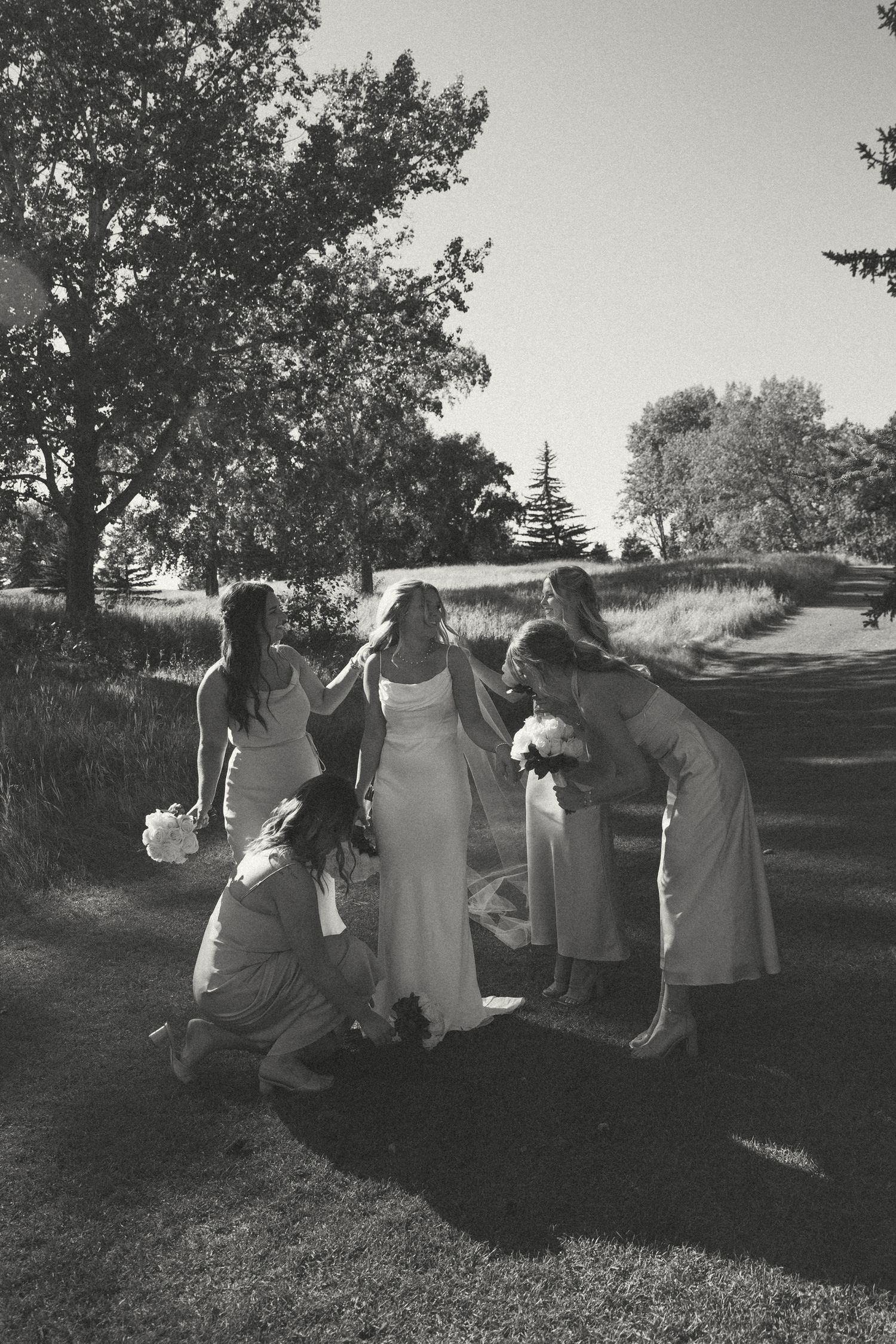 Black and white photo of bridesmaids helping adjust wedding dress outdoors.