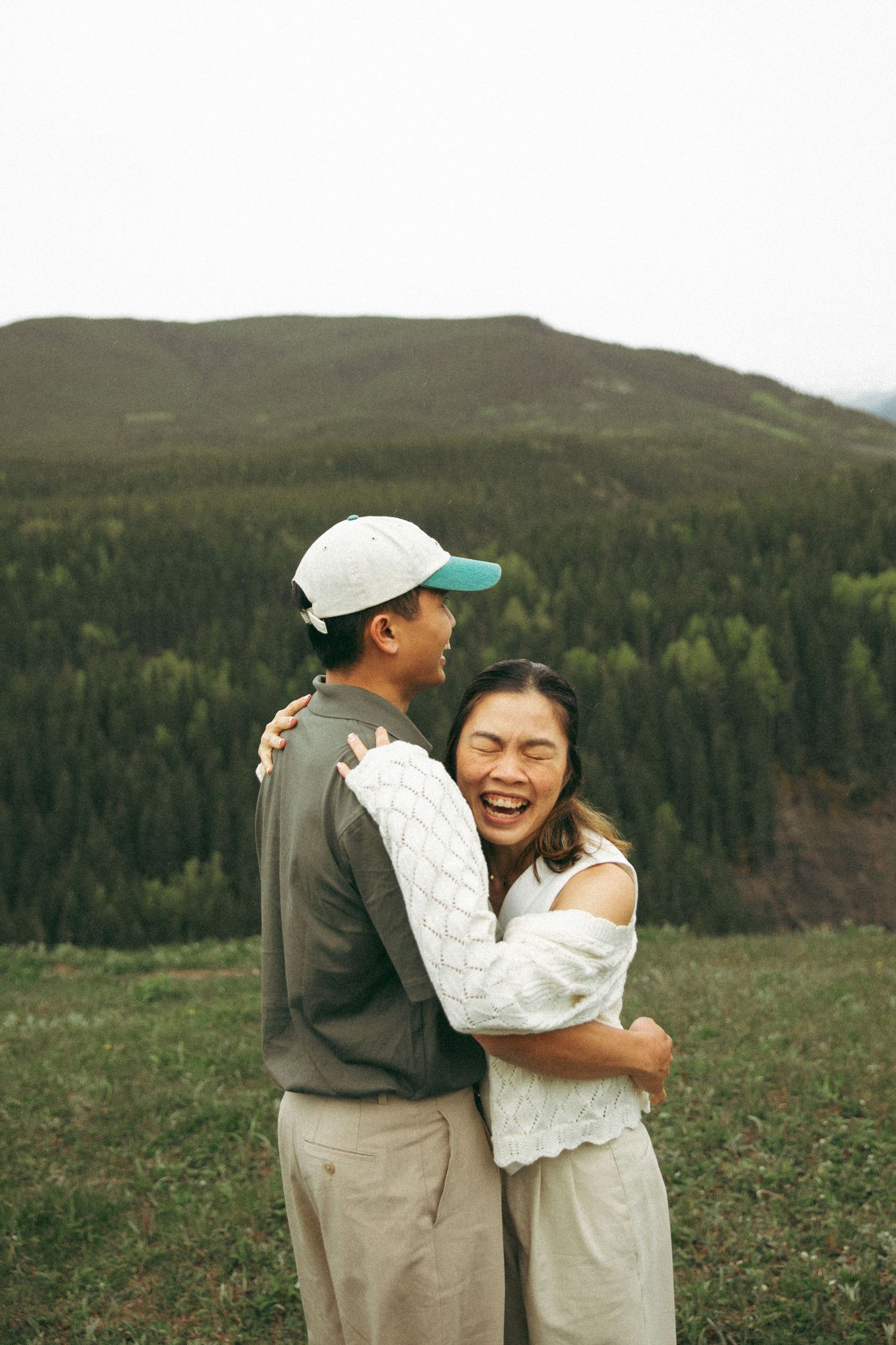 A couple embraces while enjoying a scenic mountain view on a grassy hillside.