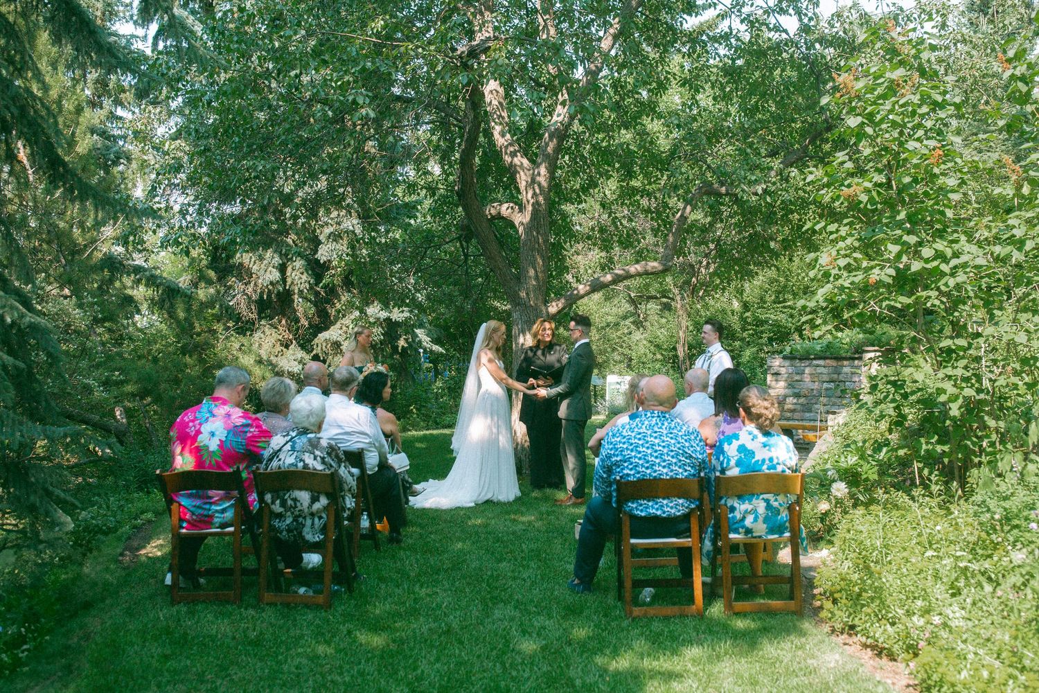 Intimate outdoor wedding ceremony with guests seated in a wooded garden setting.
