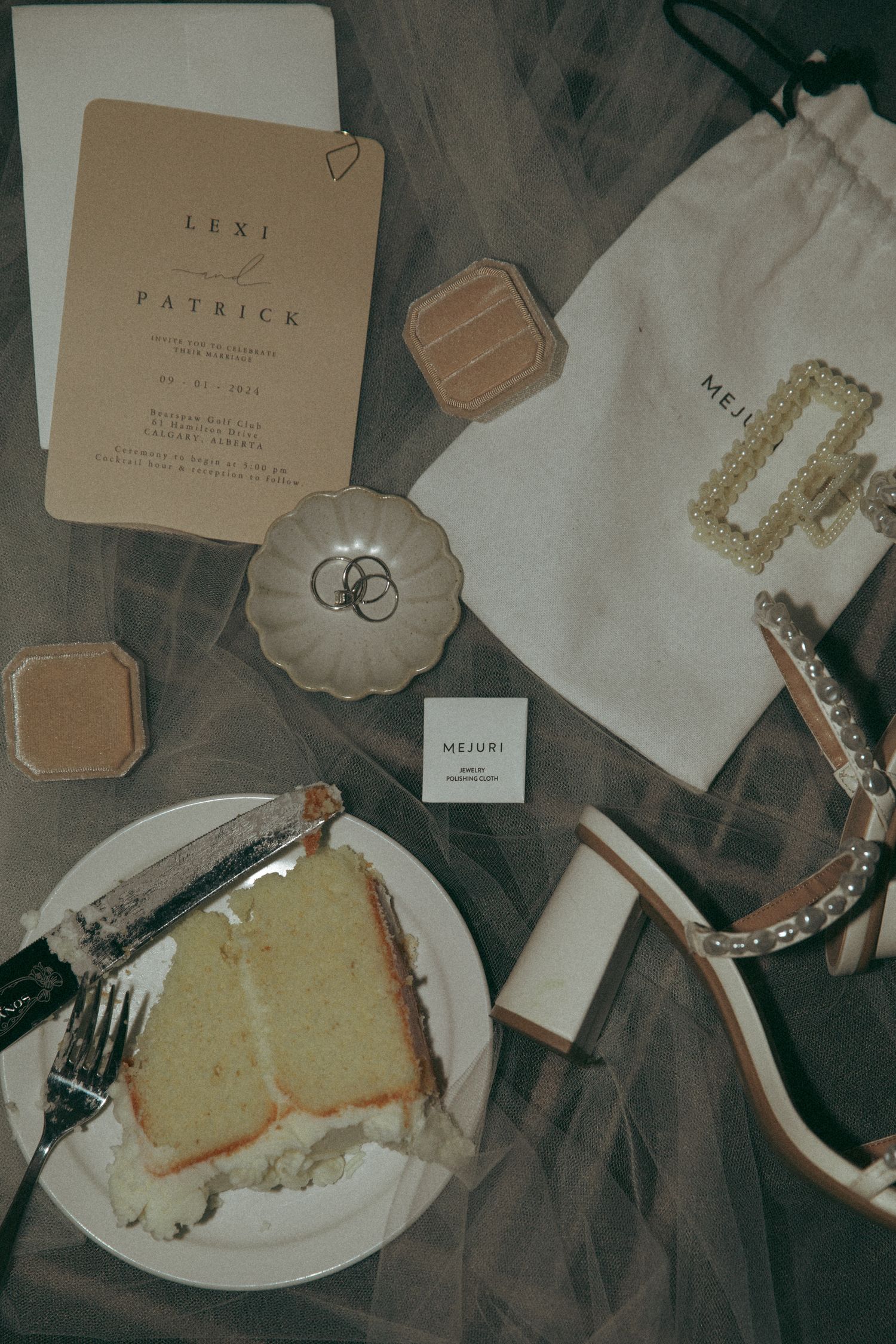 Elegant wedding details including invitation, cake slice, rings, and accessories arranged on dark surface in muted tones.