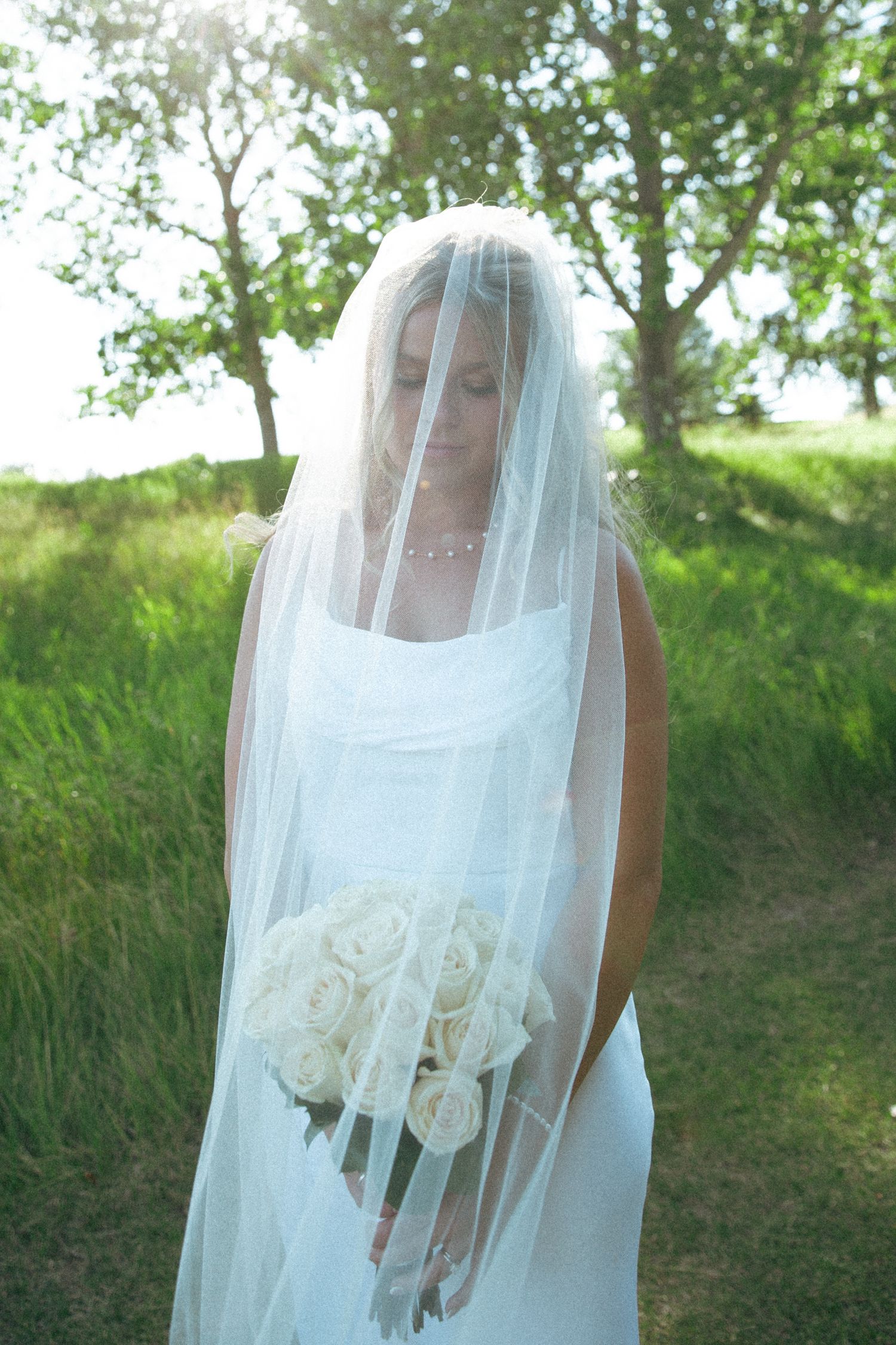 A bride in a white dress and veil holds a bouquet of white roses in an outdoor garden setting.