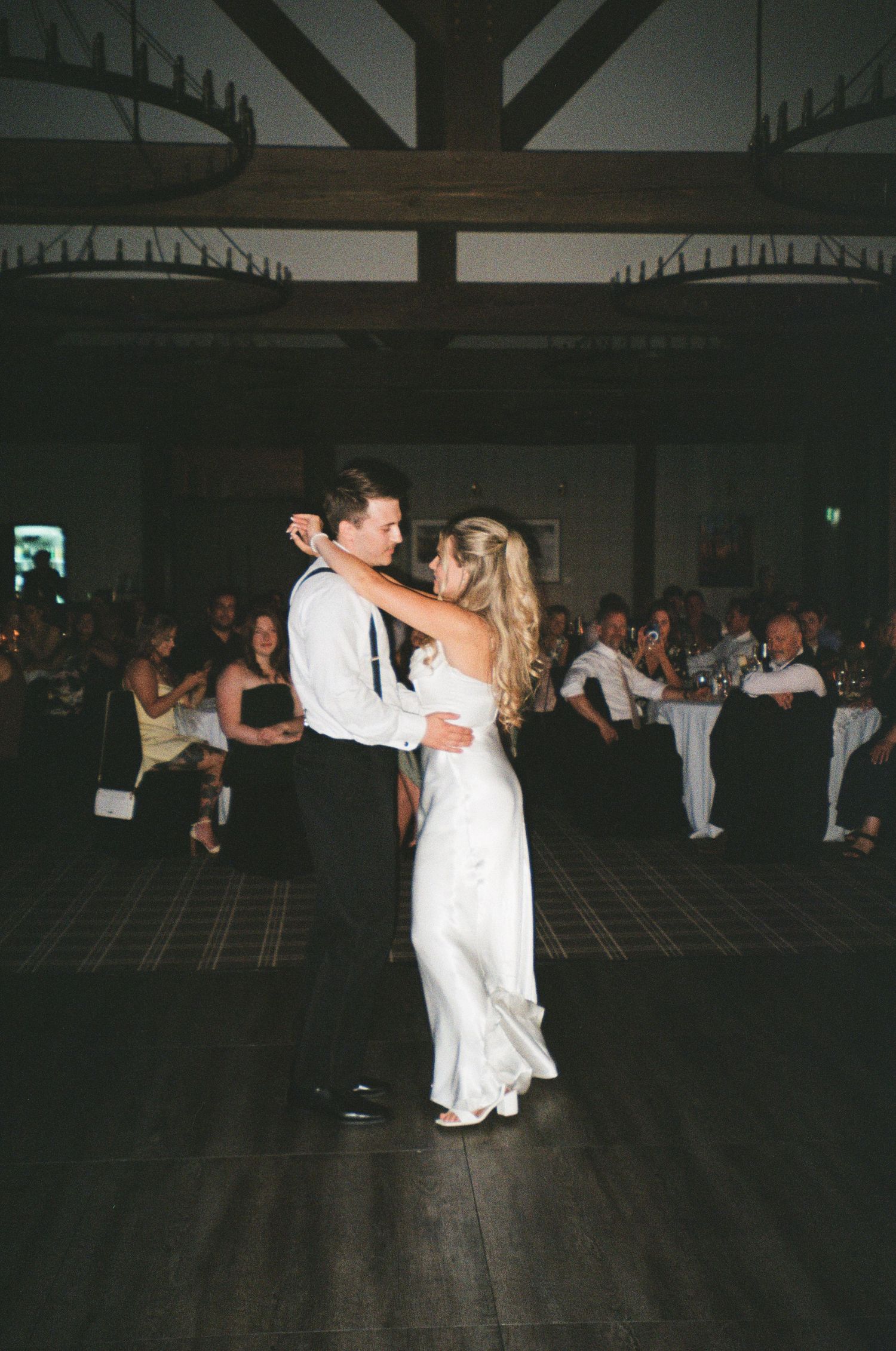 Couple shares their first dance in an elegant dark reception hall with wooden beams overhead.