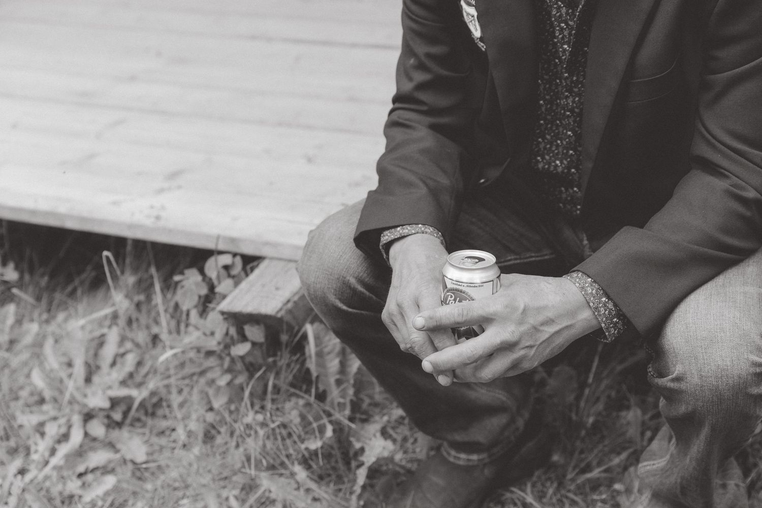 Black and white photo of a groom in a suit sitting outdoors holding a small cup at a wedding.