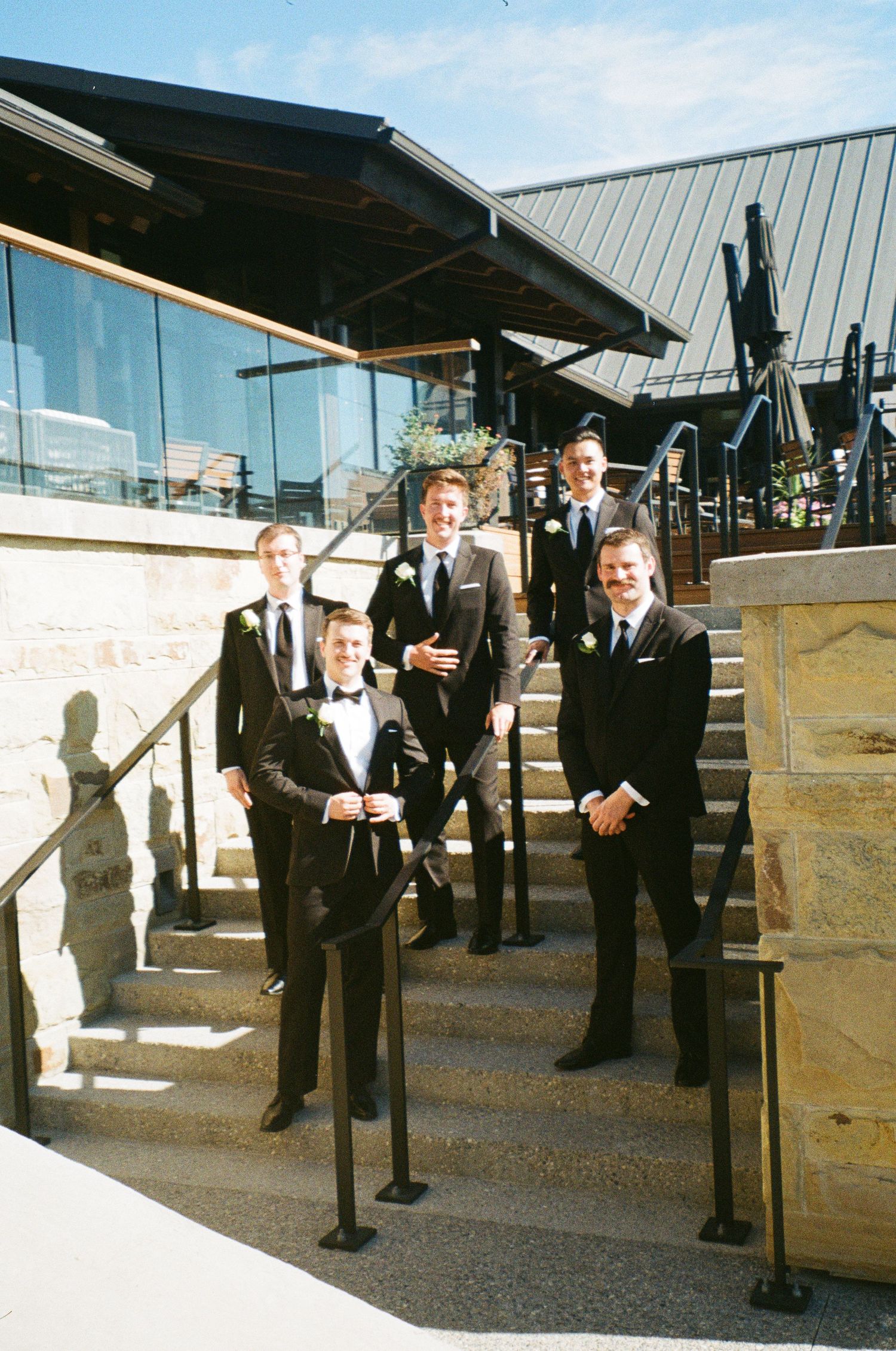 A group of formally dressed groomsmen stand on stone steps outside a modern building with glass windows.