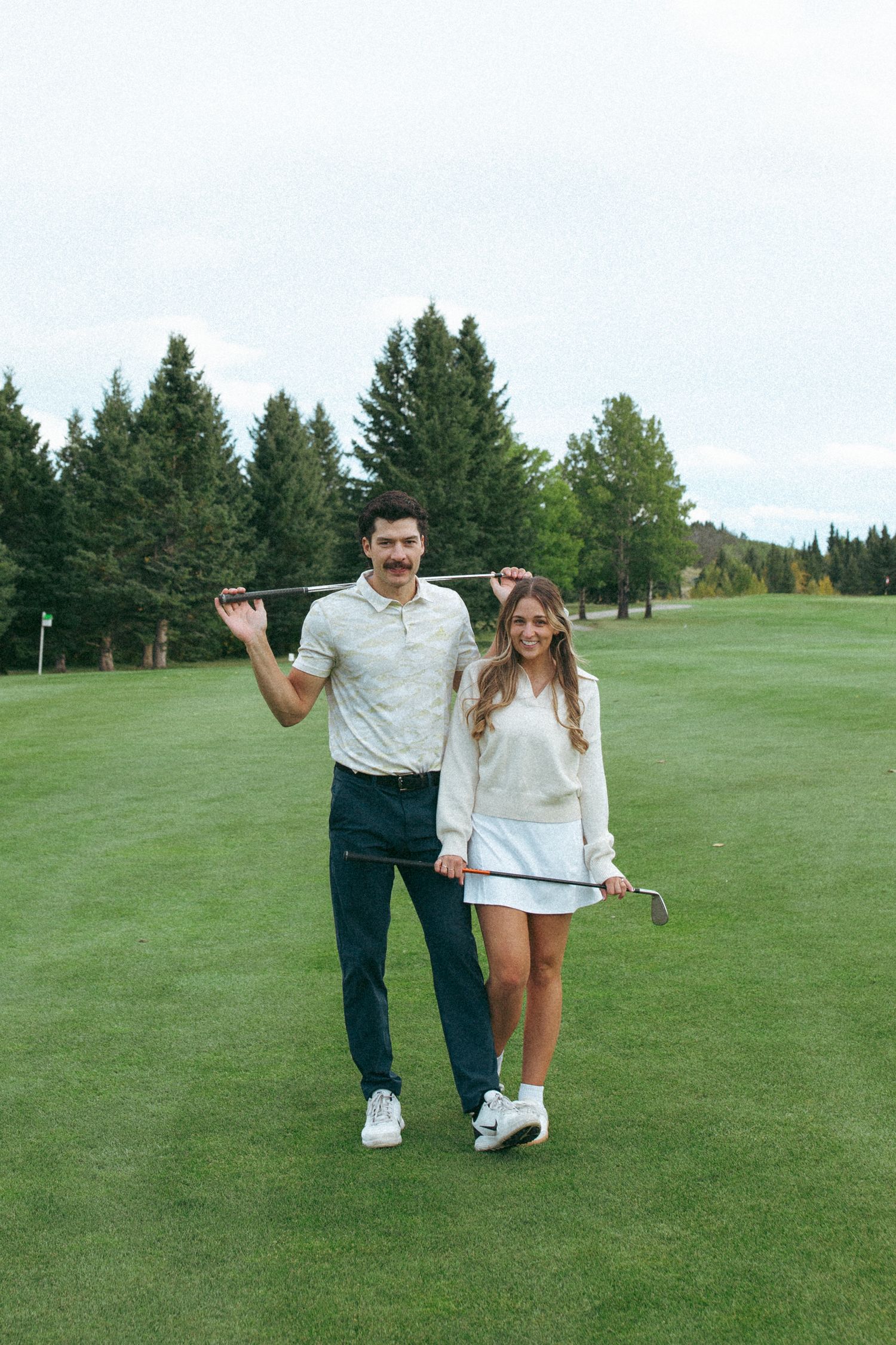 Athletic couple standing together on golf course holding clubs with evergreen forest behind them.