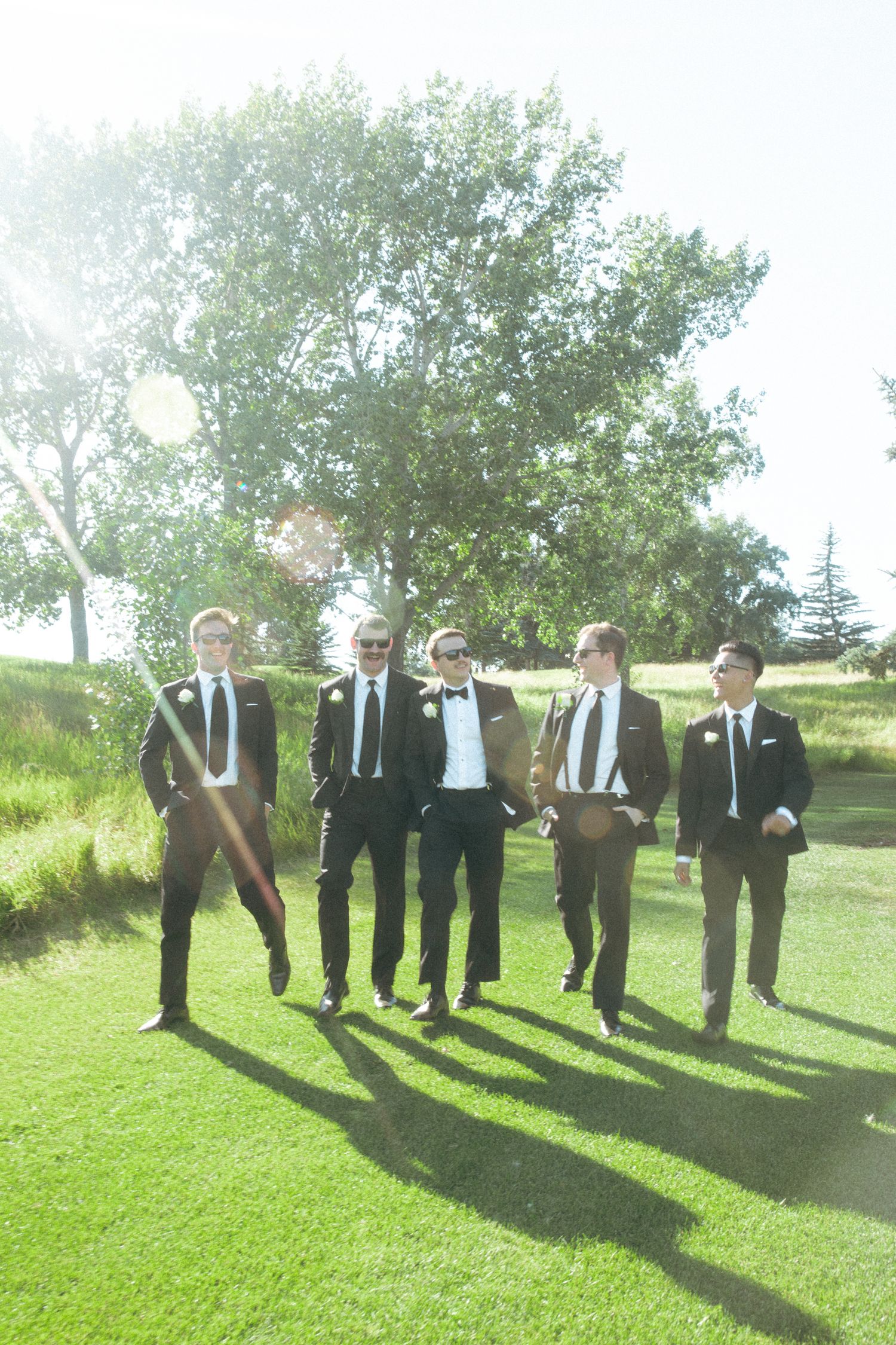 Groomsmen in dark suits pose casually on a sunny lawn with long shadows.