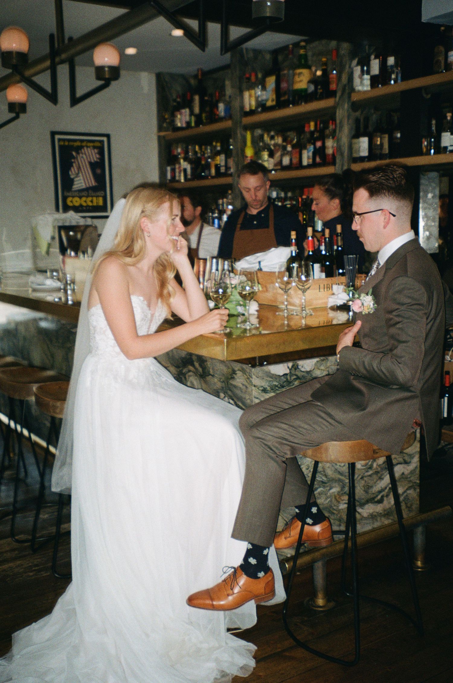 A couple in wedding attire sits at a stylish bar counter with bottles displayed on shelves in the background.