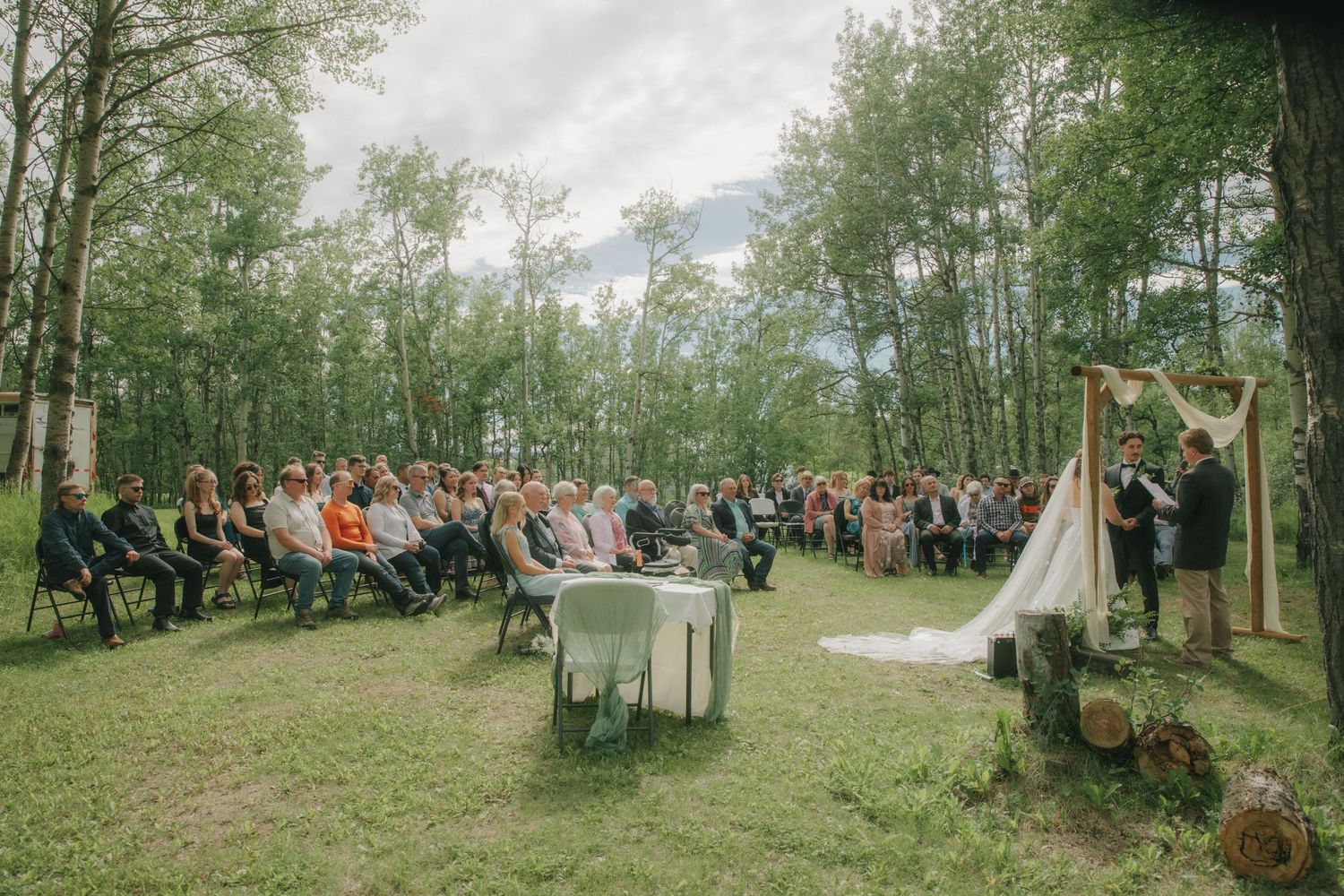 Outdoor wedding ceremony in a forest clearing with guests seated on chairs facing a floral arch altar.