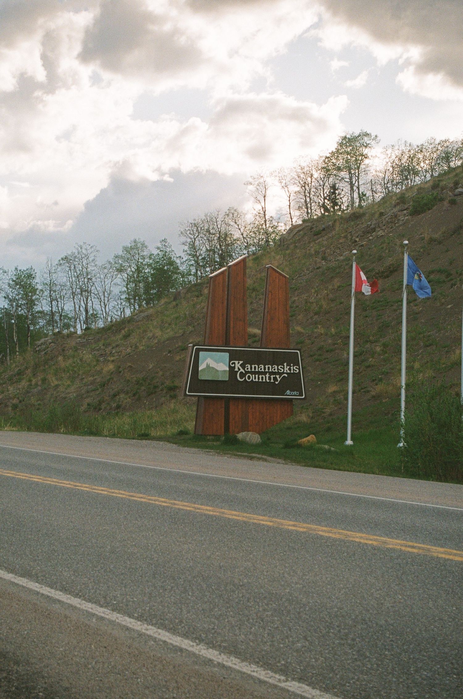 Welcome sign for 'Kananaskis Country' on hillside with two flags along mountain road.