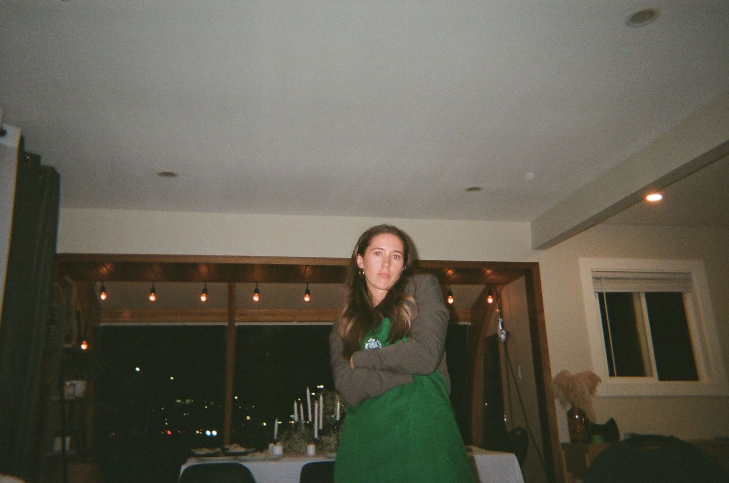 Person in a Starbucks apron standing casually in a dimly lit room with string lights in the background.