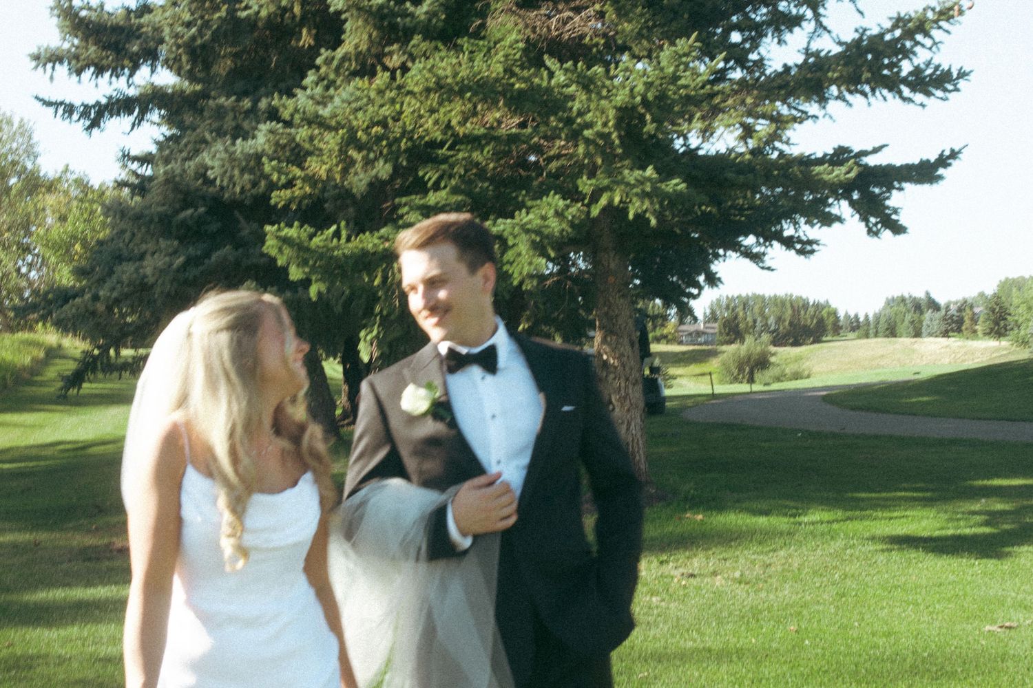 A newlywed couple shares a moment on a golf course with evergreen trees in the background.