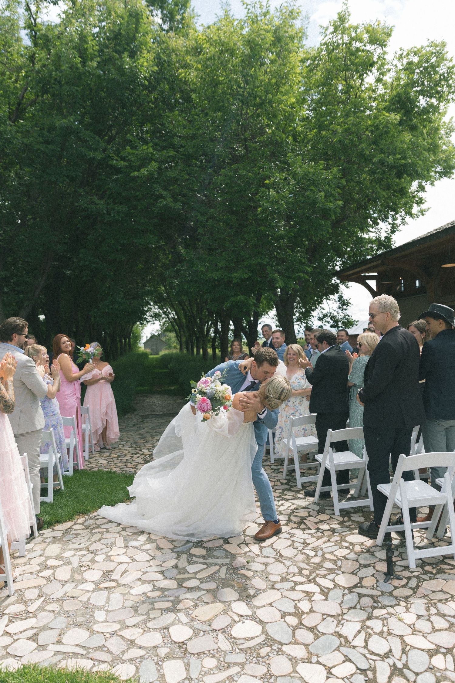 Wedding guests gather on a stone patio surrounded by lush green trees.