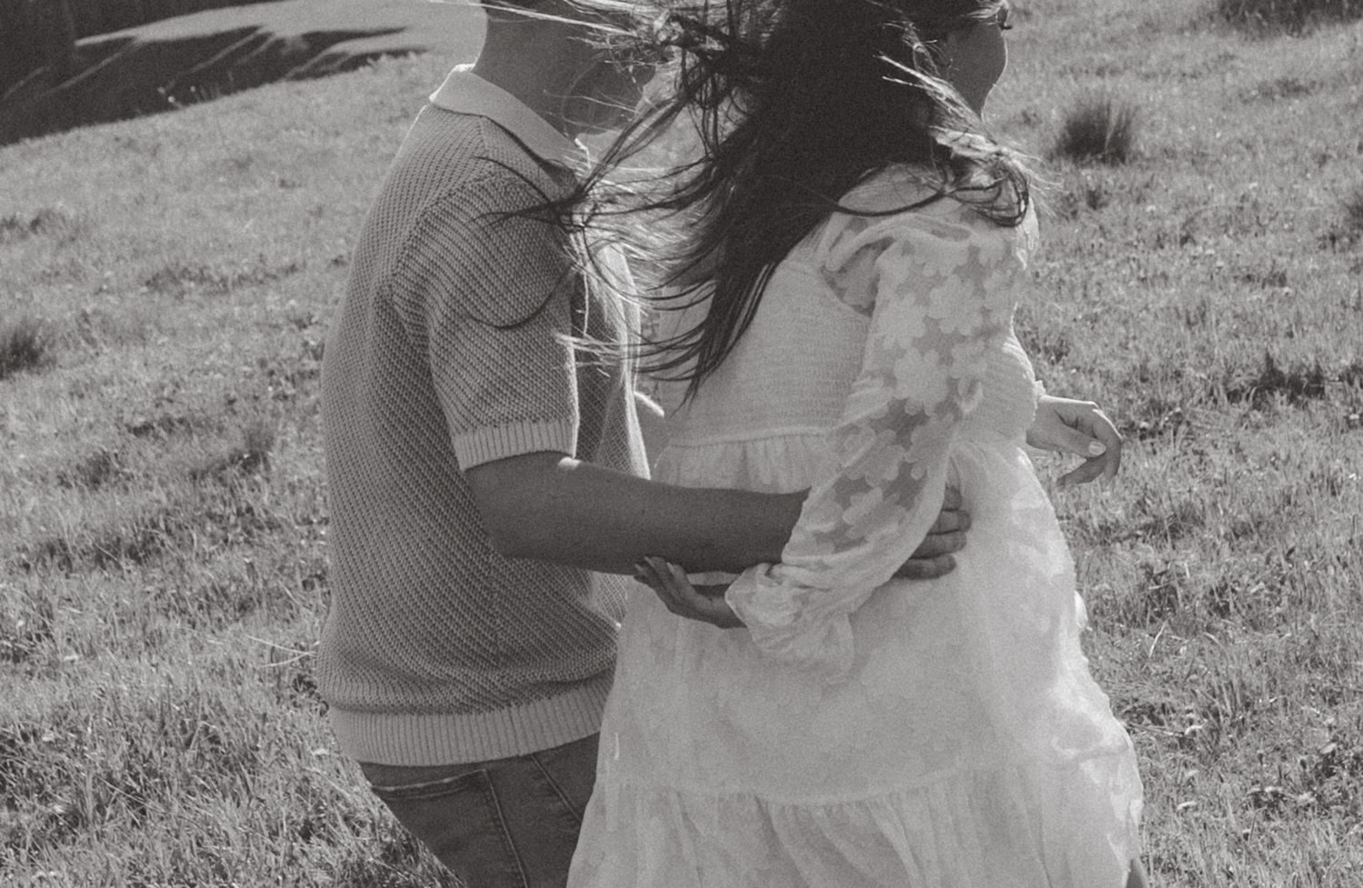 Two figures in black and white photograph embrace while spinning in a grassy field on a windy day.