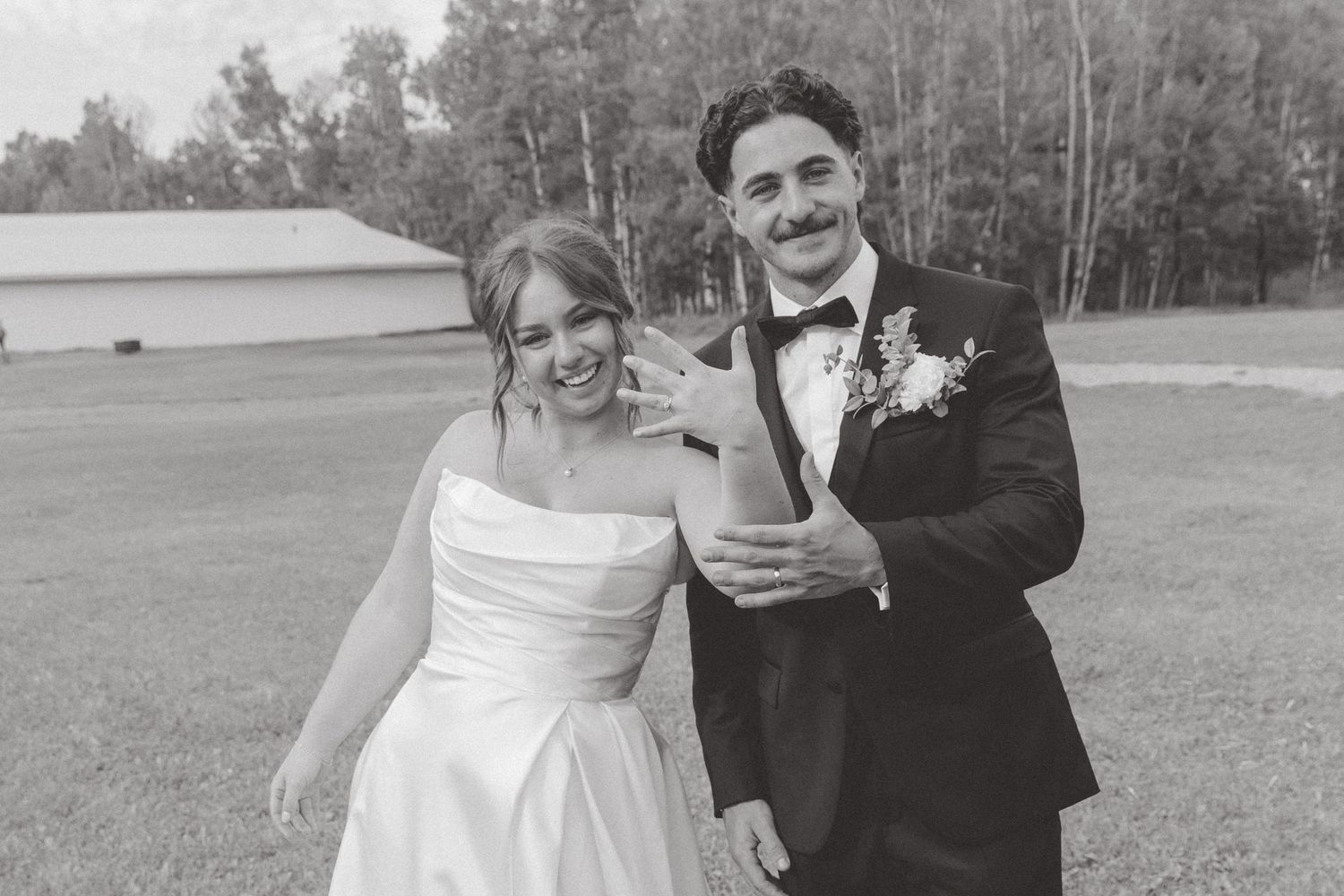 Black and white photo of a smiling bride in strapless gown and groom in tuxedo with boutonniere on their wedding day.
