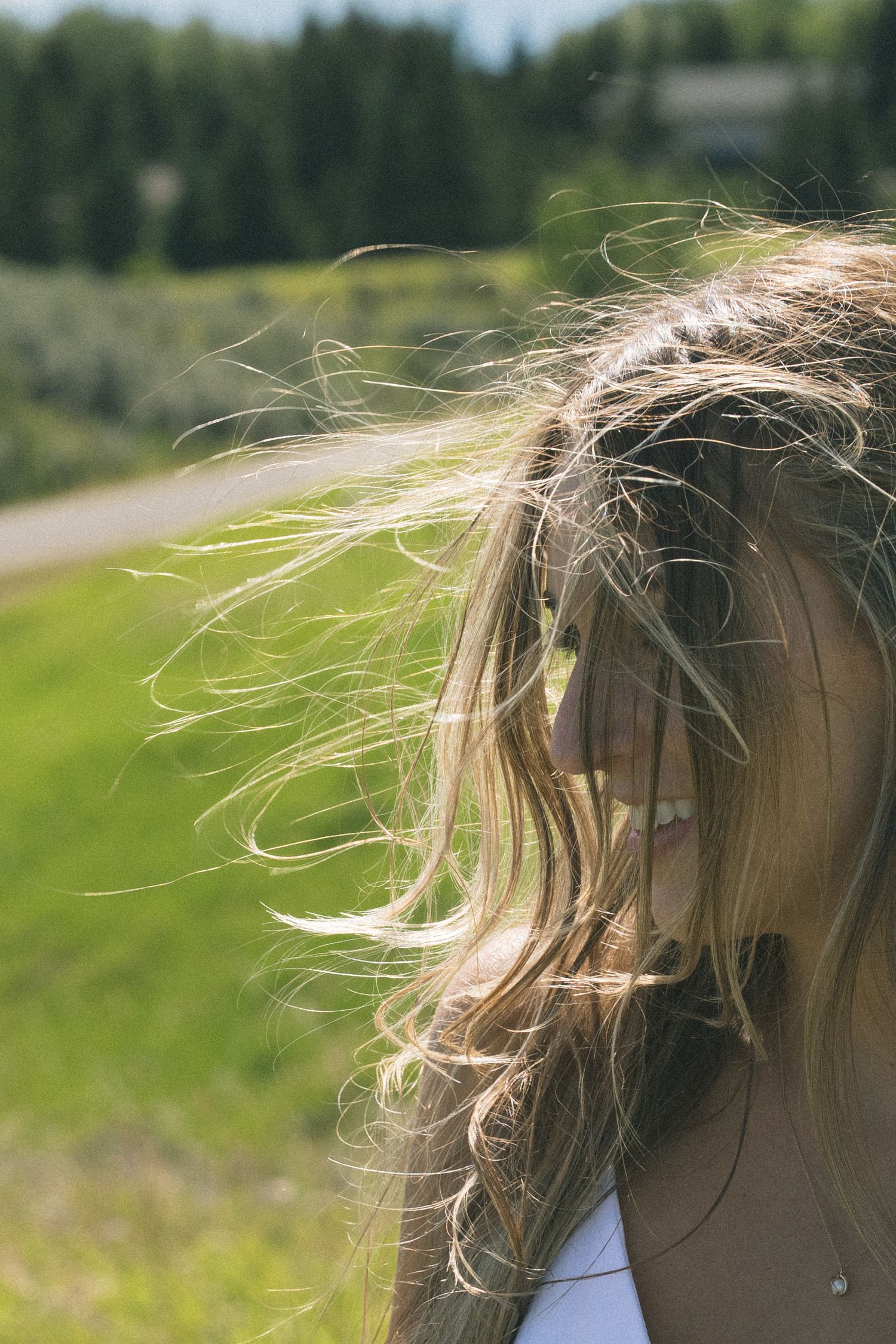 Blonde woman smiling outdoors with wind-blown hair against a blurred green landscape background.